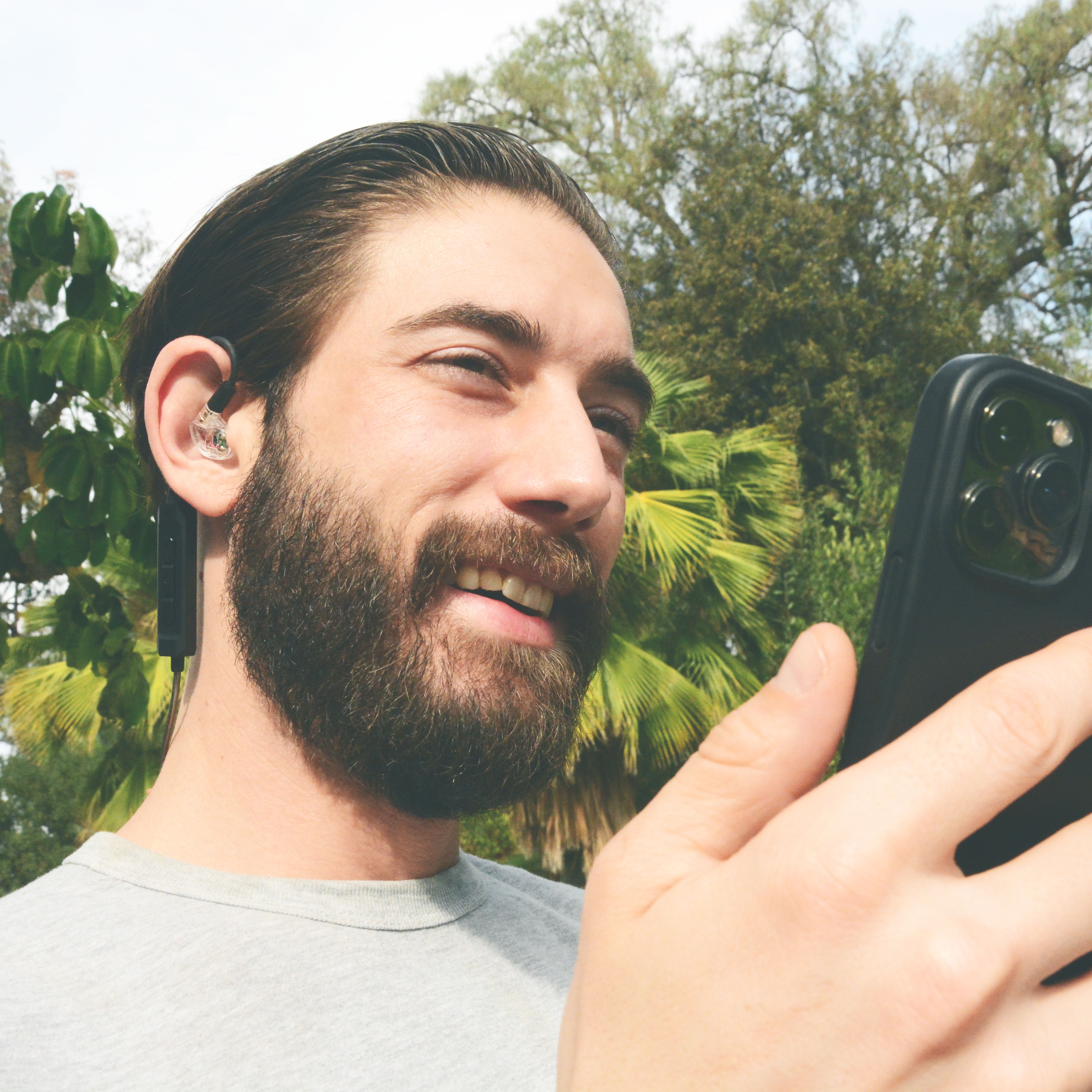 Man with a beard wearing the MEE audio M6 PRO Musicians' In-Ear Monitors Wired + Wireless Combo Pack: Includes IEMs, 2 Audio Cables and BT3 Hi-Res Bluetooth Audio Adapter w/aptX, LDAC, Auracast Support, & Built-In Headset (Clear) while using a smartphone outdoors with greenery in the background