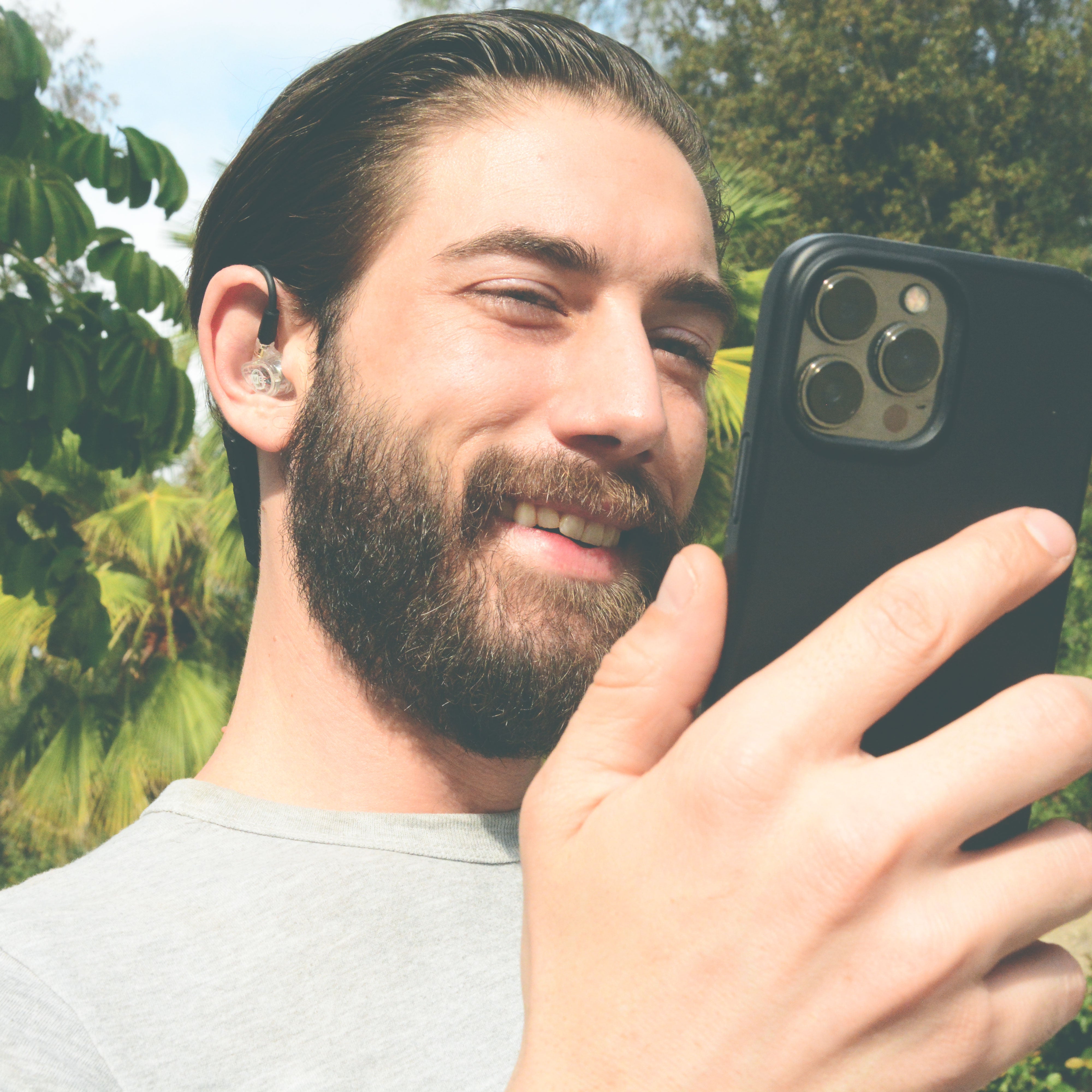 Man with a beard wearing the MEE audio MX1 PRO Musicians' In-Ear Monitors Wired + Wireless Combo Pack: Includes MX1 PRO Dynamic Driver IEMs, 2 Cables, and BT3 Hi-Res Bluetooth Audio Adapter w/aptX, LDAC & Built-in Headset (Clear) holding a smartphone outdoors with greenery in the background