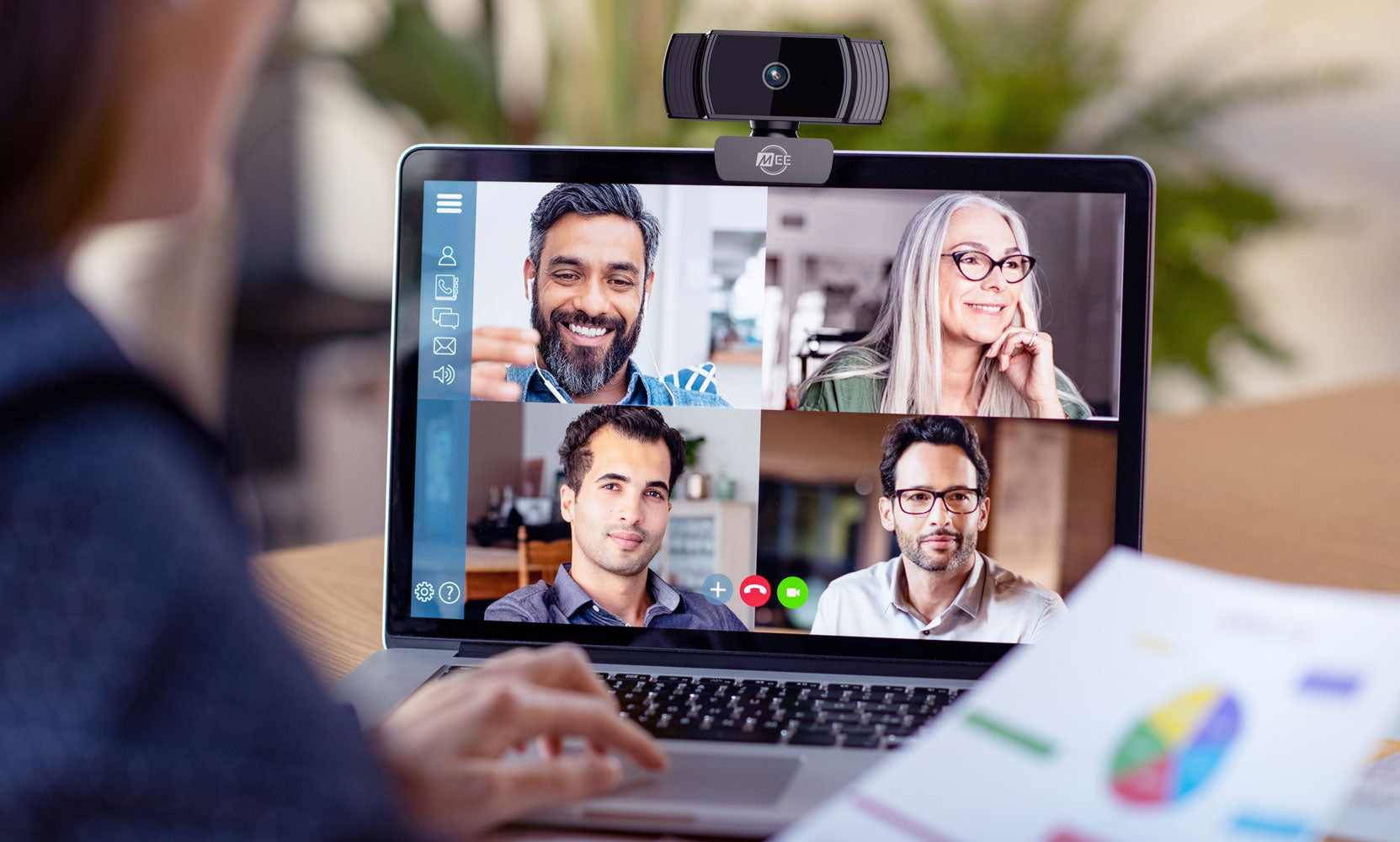 An over-the-shoulder view of a woman's laptop screen showing a video conference with four participants, each in separate frames, smiling and engaging in discussion.
