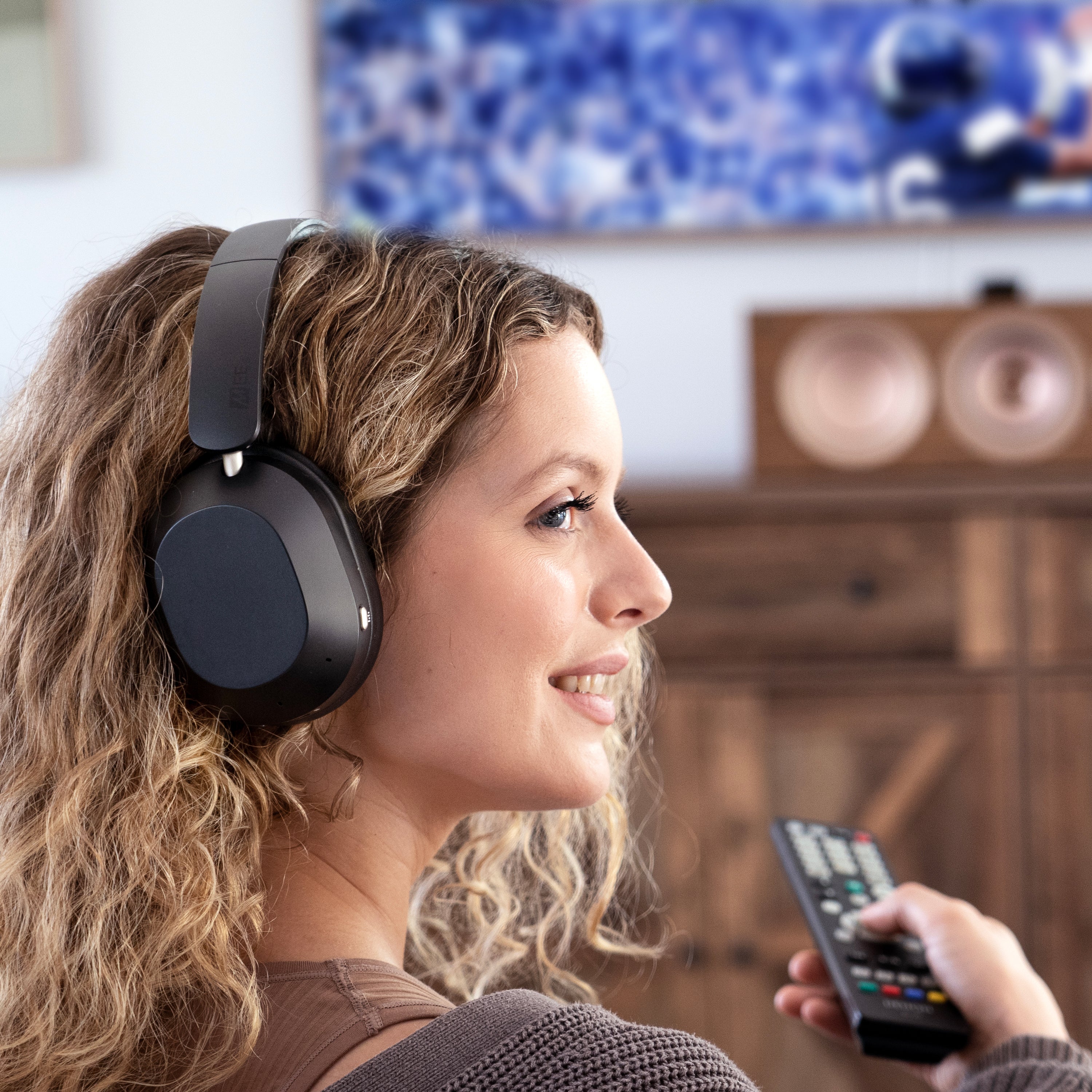 Woman wearing MEE audio Matrix Cinema 3D 2026 Bluetooth Headphones + Connect Bluetooth Audio Transmitter Bundle for All TVs and holding a remote control, with a blurred background of a television and wooden cabinet.