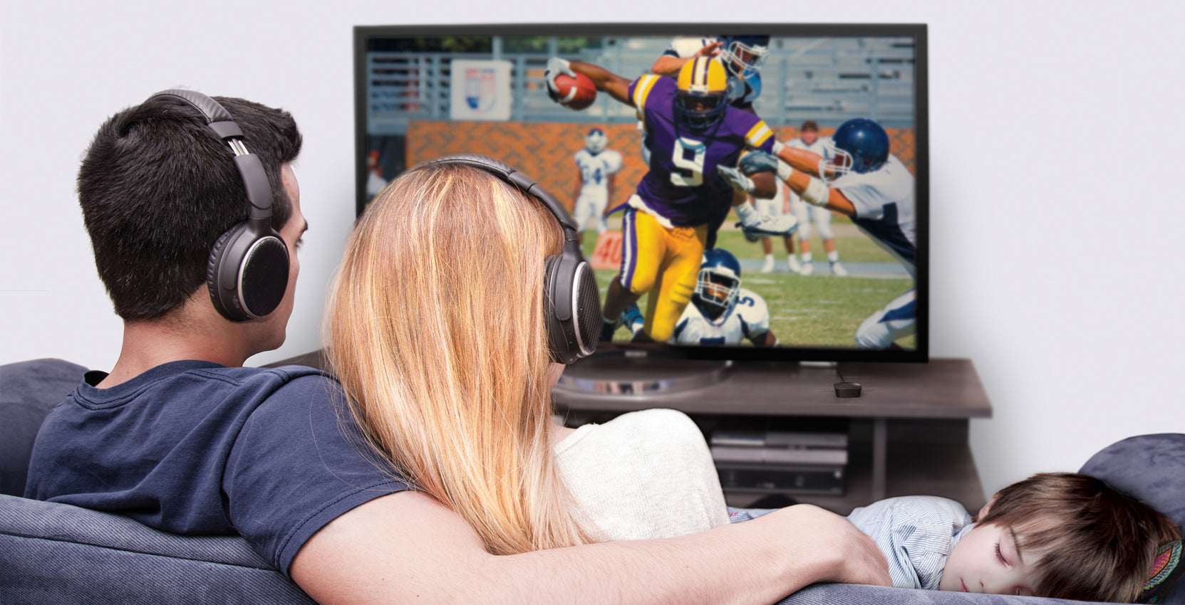 A couple wearing headphones watches a football game on tv while their child sleeps on the couch beside them.
