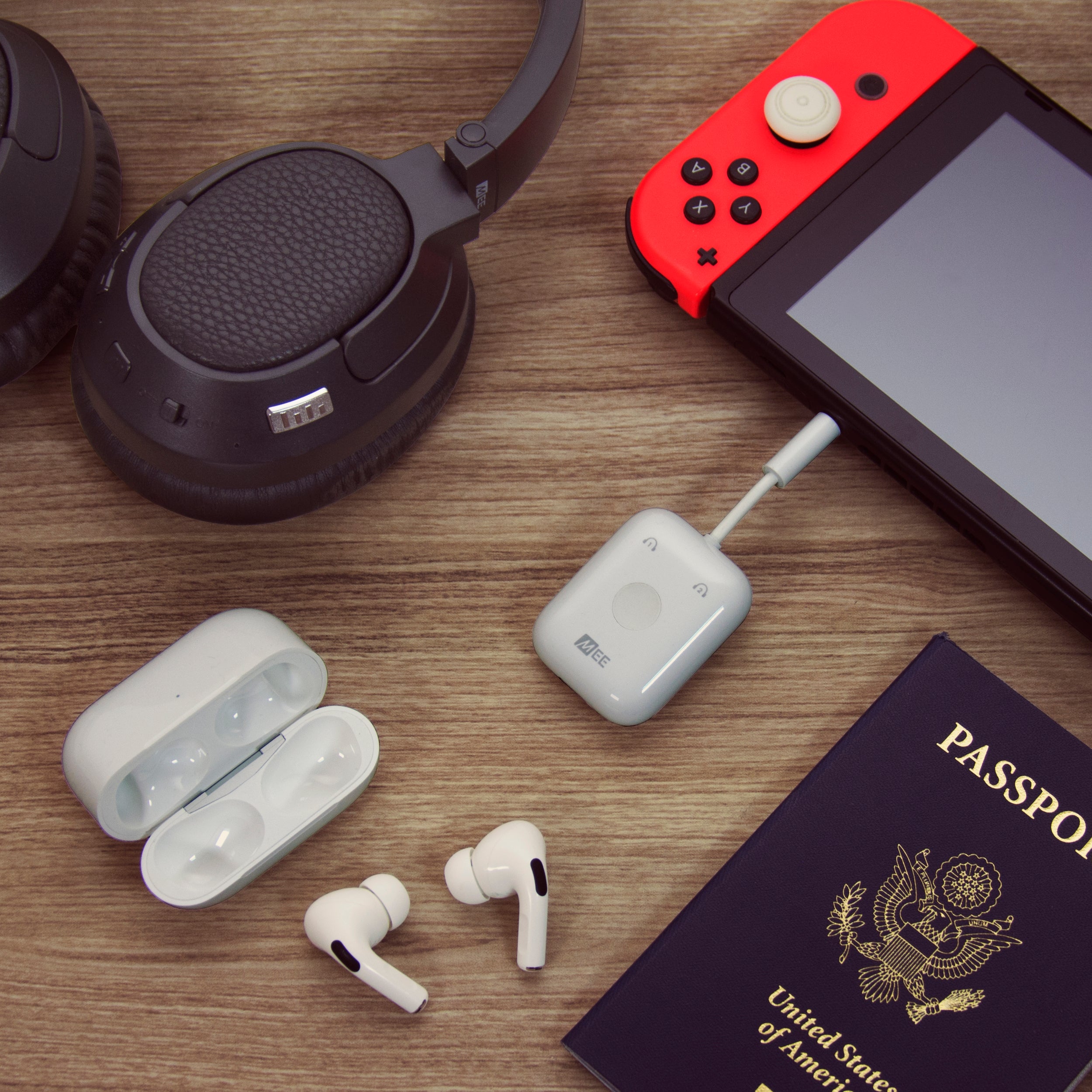 A flat lay photo featuring a passport, headphones, a handheld gaming console, a wireless transmitter bluetooth travel adapters, and earbuds on a wooden surface.