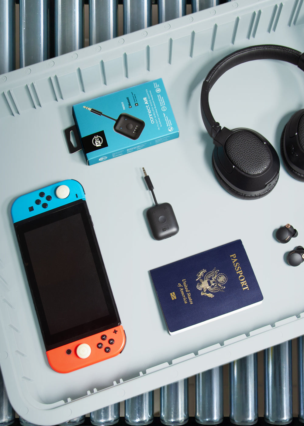A neatly arranged travel pack on a ridged surface with a blue and red game console, a pair of headphones, an bluetooth travel adapters, earbuds, and a u.s. passport.
