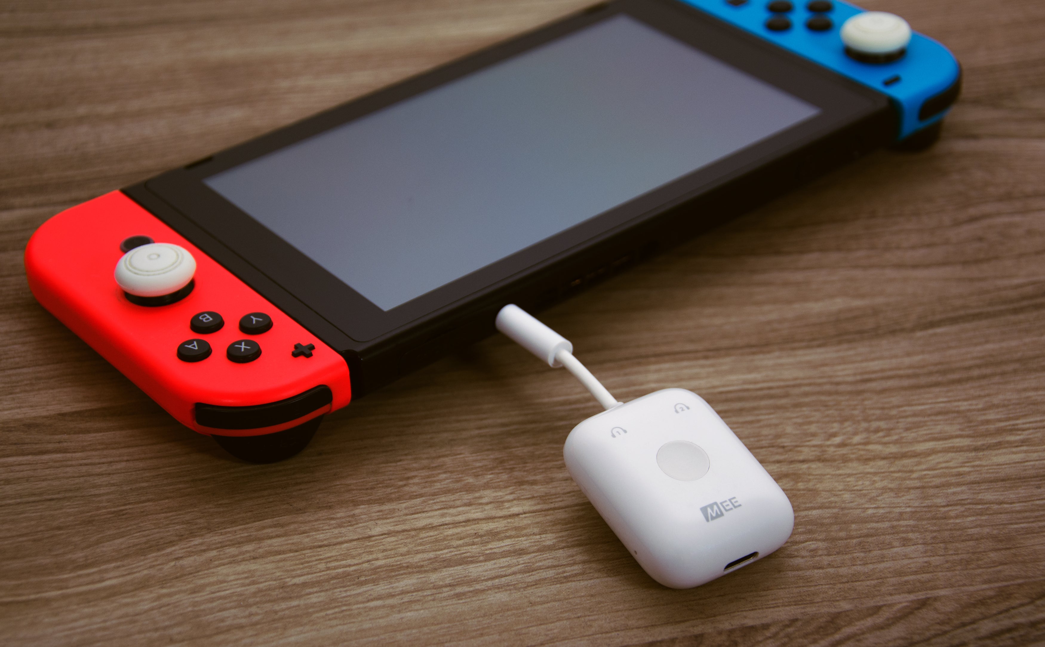 A nintendo switch with red and blue controllers connected, lying on a wooden surface next to a white usb-c audio transmitter.