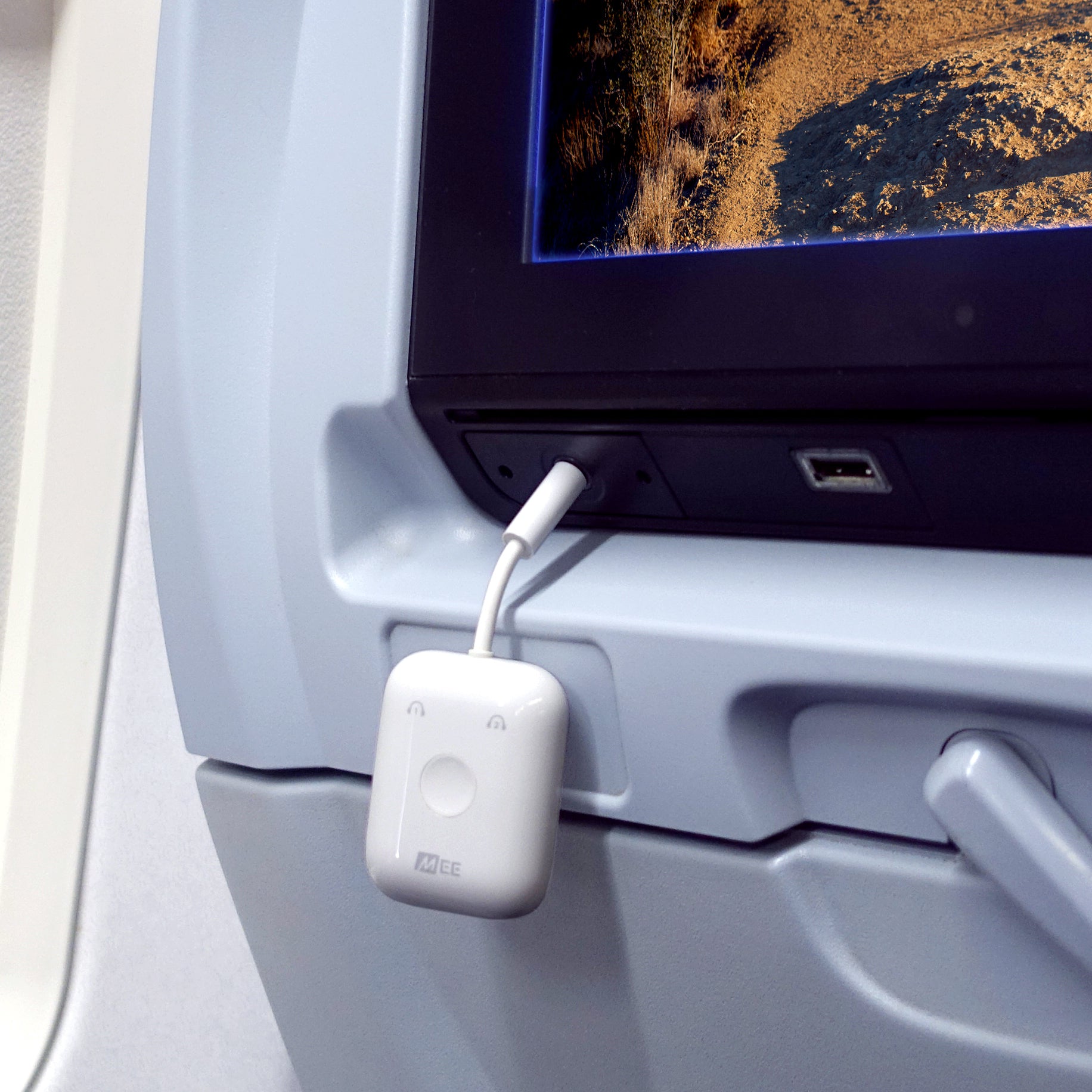 A white portable Bluetooth travel adapters device plugged into an airplane seat's usb port, charging a connected gadget. the screen above displays a view of earth’s surface from above.
