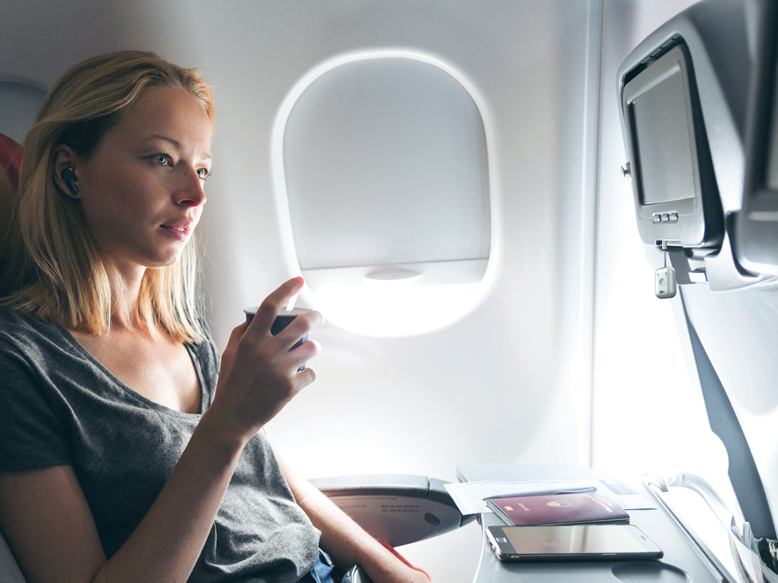 A woman sitting by the window in an airplane, looking absorbed in her bluetooth travel adapters with flight entertainment system beside and passport on her tray table.