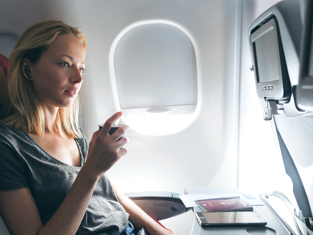 A woman sitting by the window in an airplane, looking absorbed in her smartphone with flight entertainment system beside and passport on her tray table.