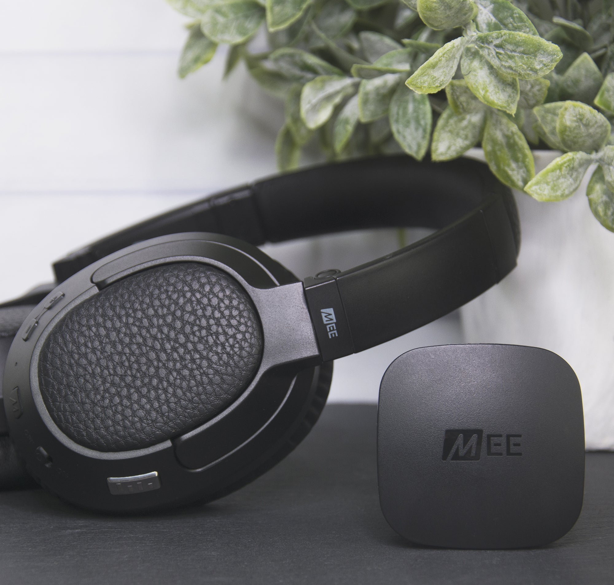 Black over-ear headphones with textured ear cups next to a small square wireless transmitter, both branded with "mee". positioned on a table beside a potted plant with green and white leaves, against a white wooden background.