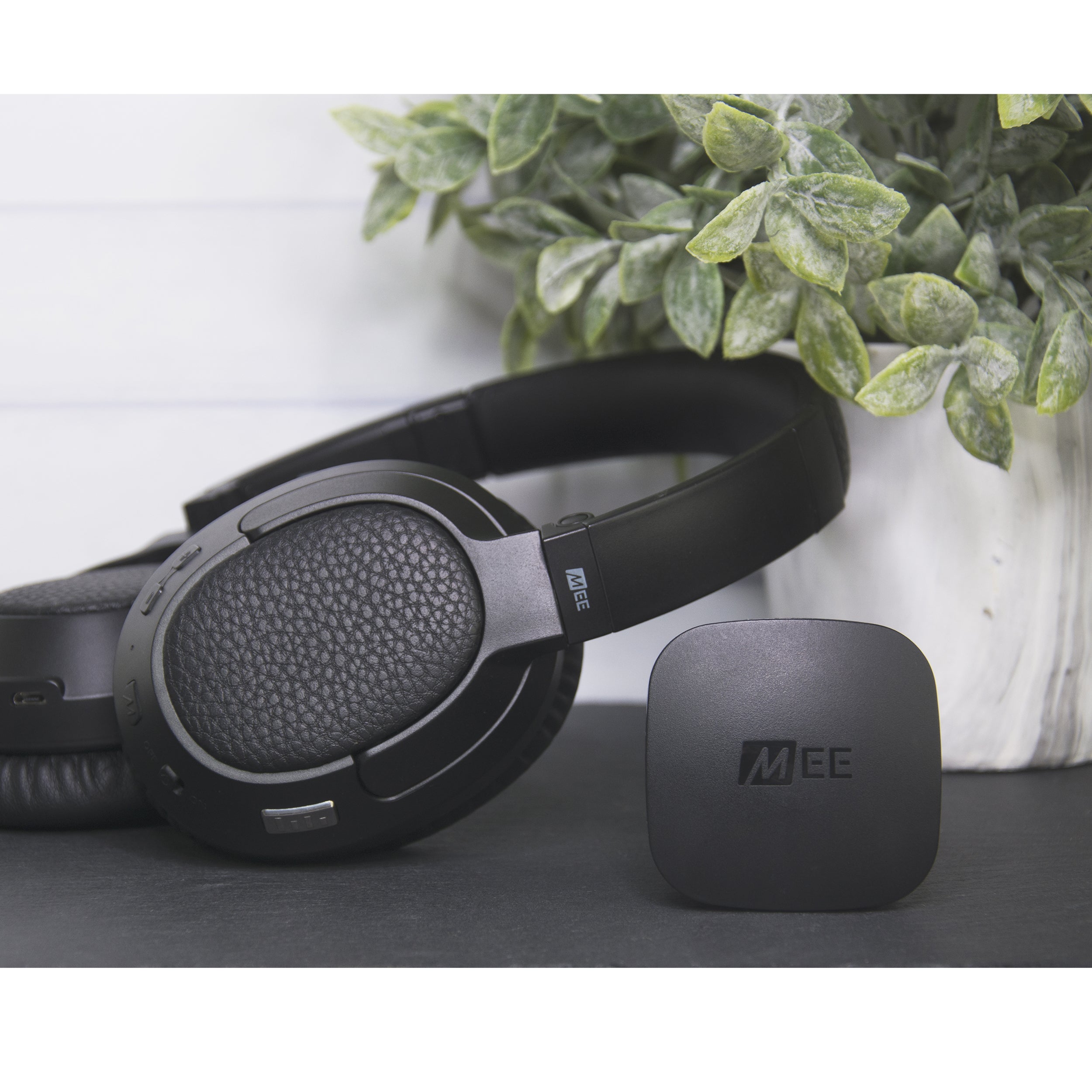 Black over-ear headphones with textured ear cups beside a small black device labeled "mee," placed on a wooden surface near a potted green plant with white background.