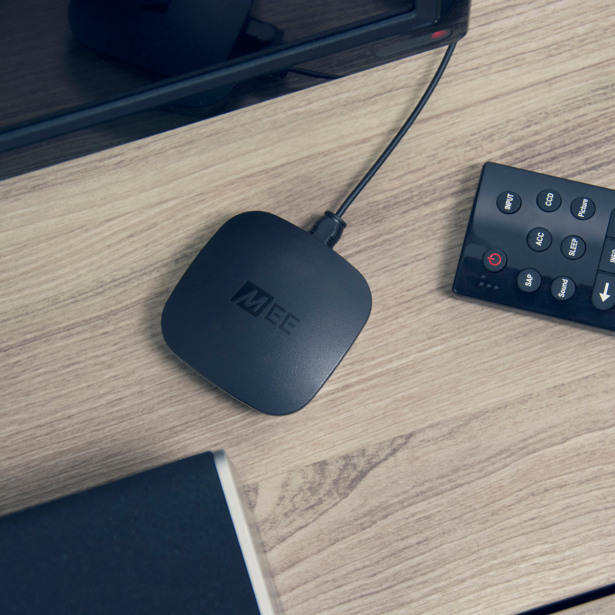A black streaming media player labeled "mee" connected to a cable on a wooden desk, next to a remote control and a part of a black electronic device.