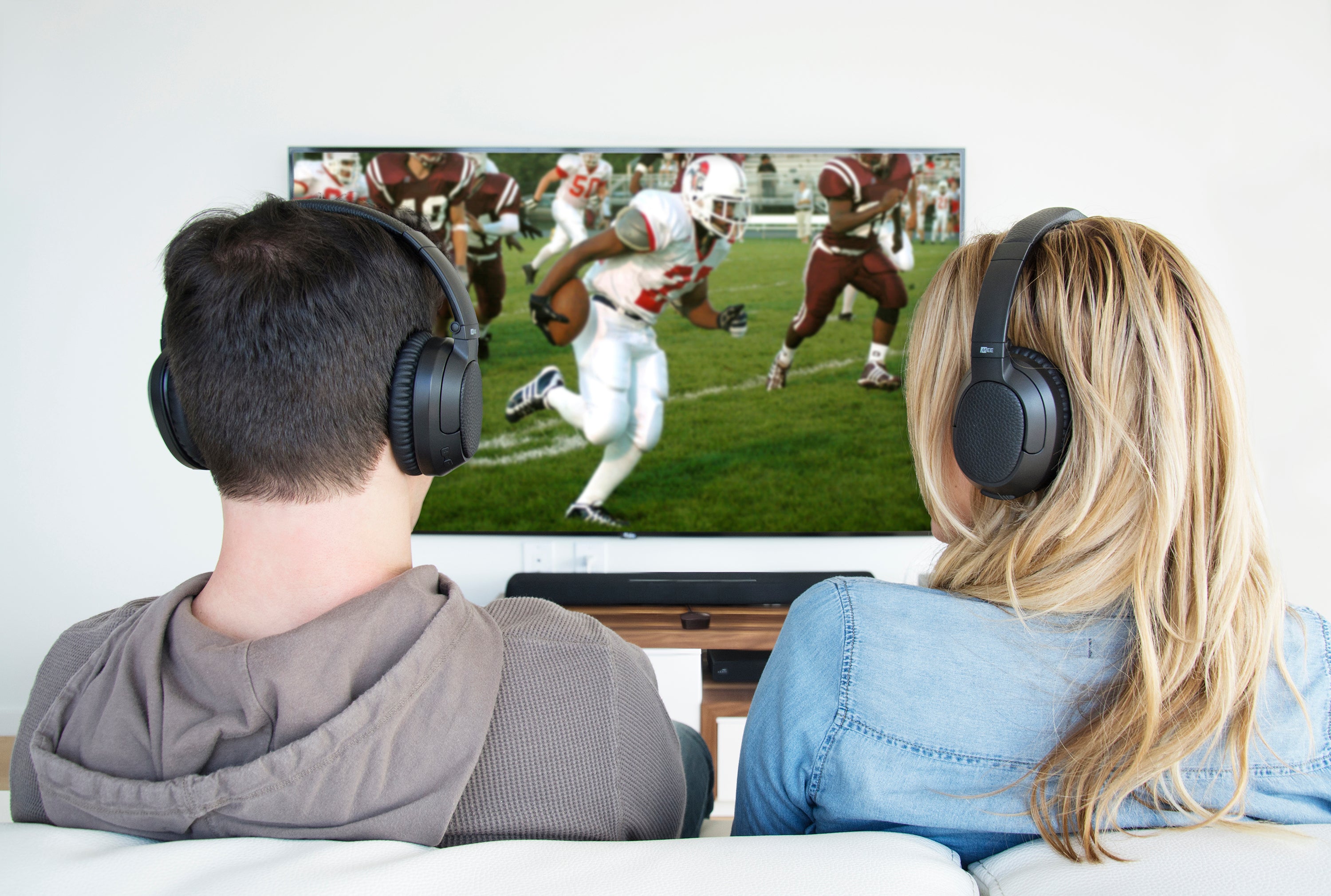 A man and a woman wearing headphones sit on a couch, watching a football game on a large tv screen in a bright living room.