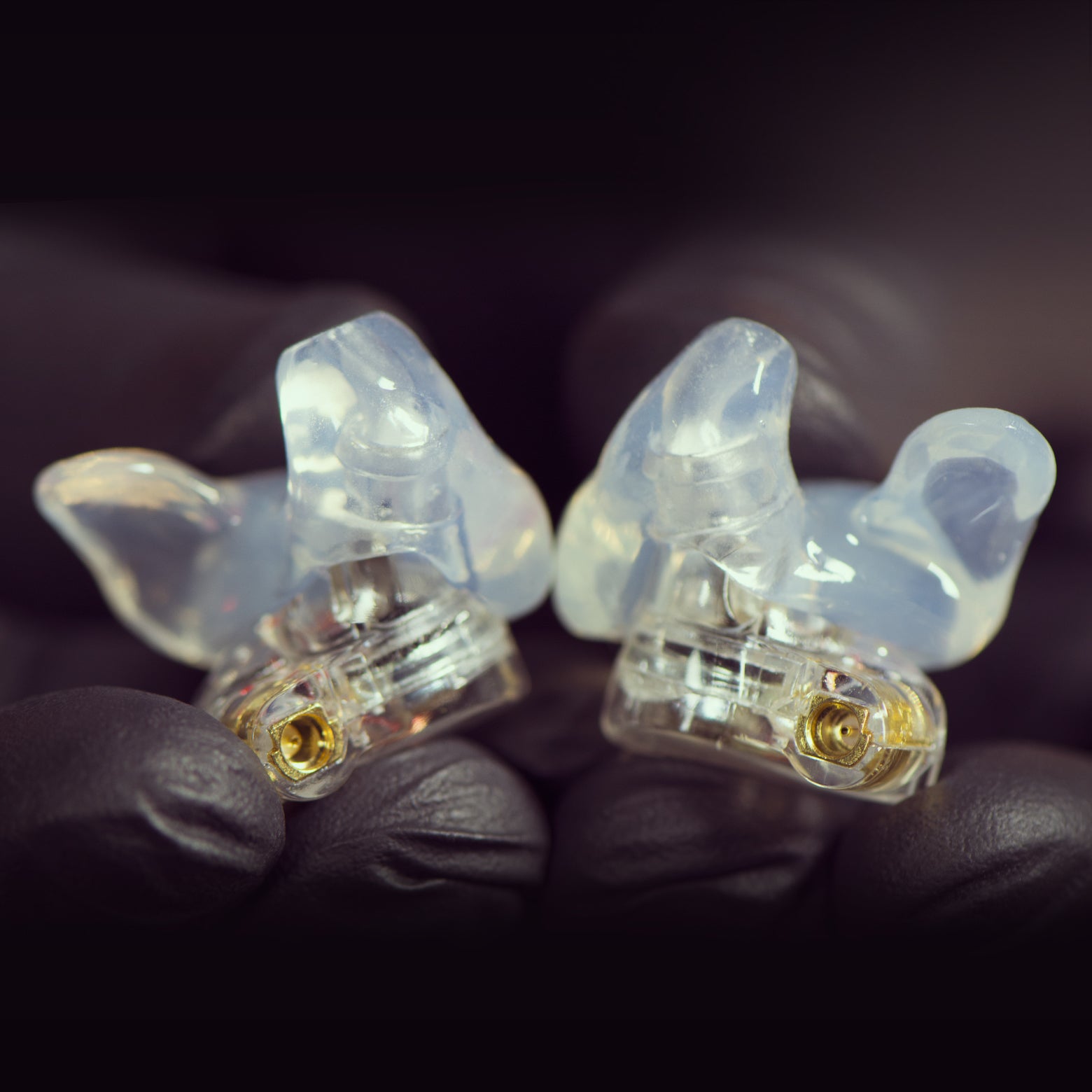 Two custom-molded, clear in-ear monitors held carefully between fingertips against a dark background, focusing on their detailed, translucent structure.