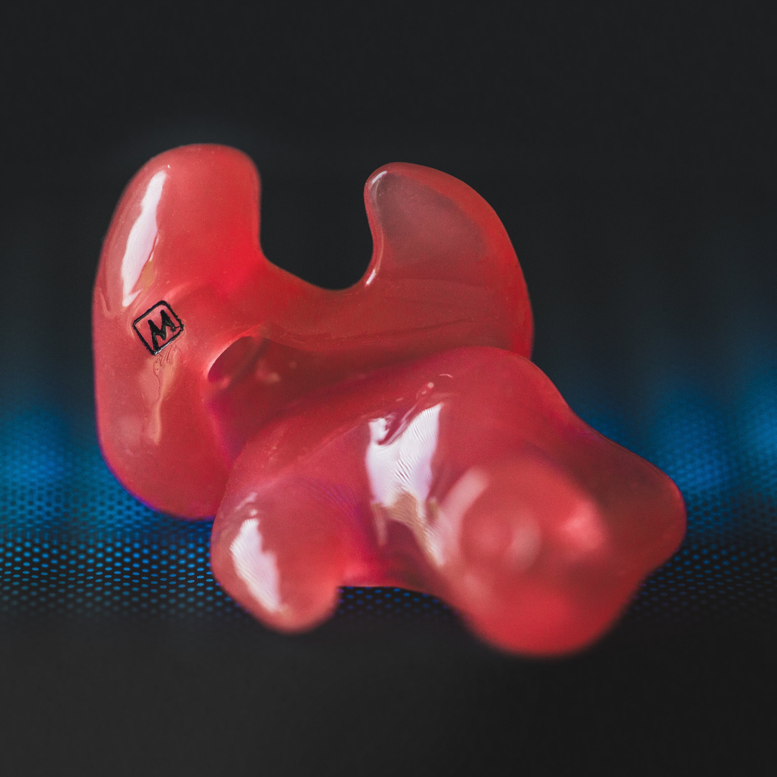 Close-up of a red, custom-made earplug with the letter "m" marked on it, placed on a dark blue textured surface.
