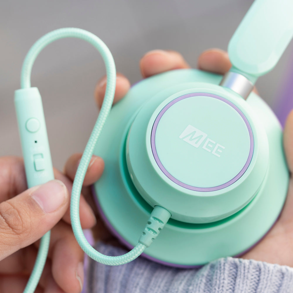 A close-up of a hand holding mint KJ45 KidJamz headphones with a textured cable. the headphone has a visible brand logo "MEE" in the center.