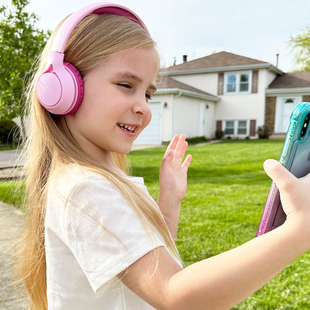 A young girl wearing pink headphones smiles and waves at a smartphone she is holding in her hand, standing outdoors on a lawn with a house in the background.