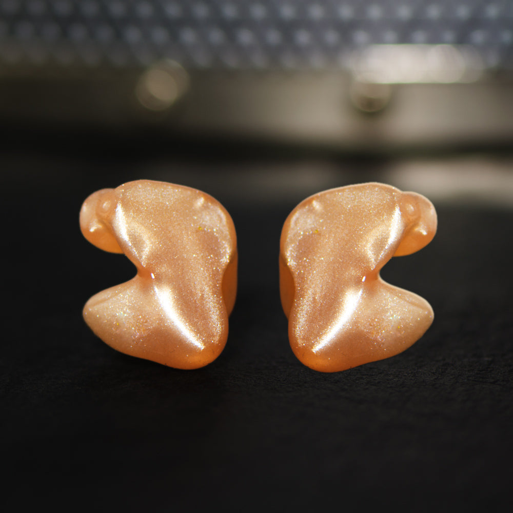 Two sparkly peach-colored custom earplugs on a dark, textured surface, highlighted by soft natural light.