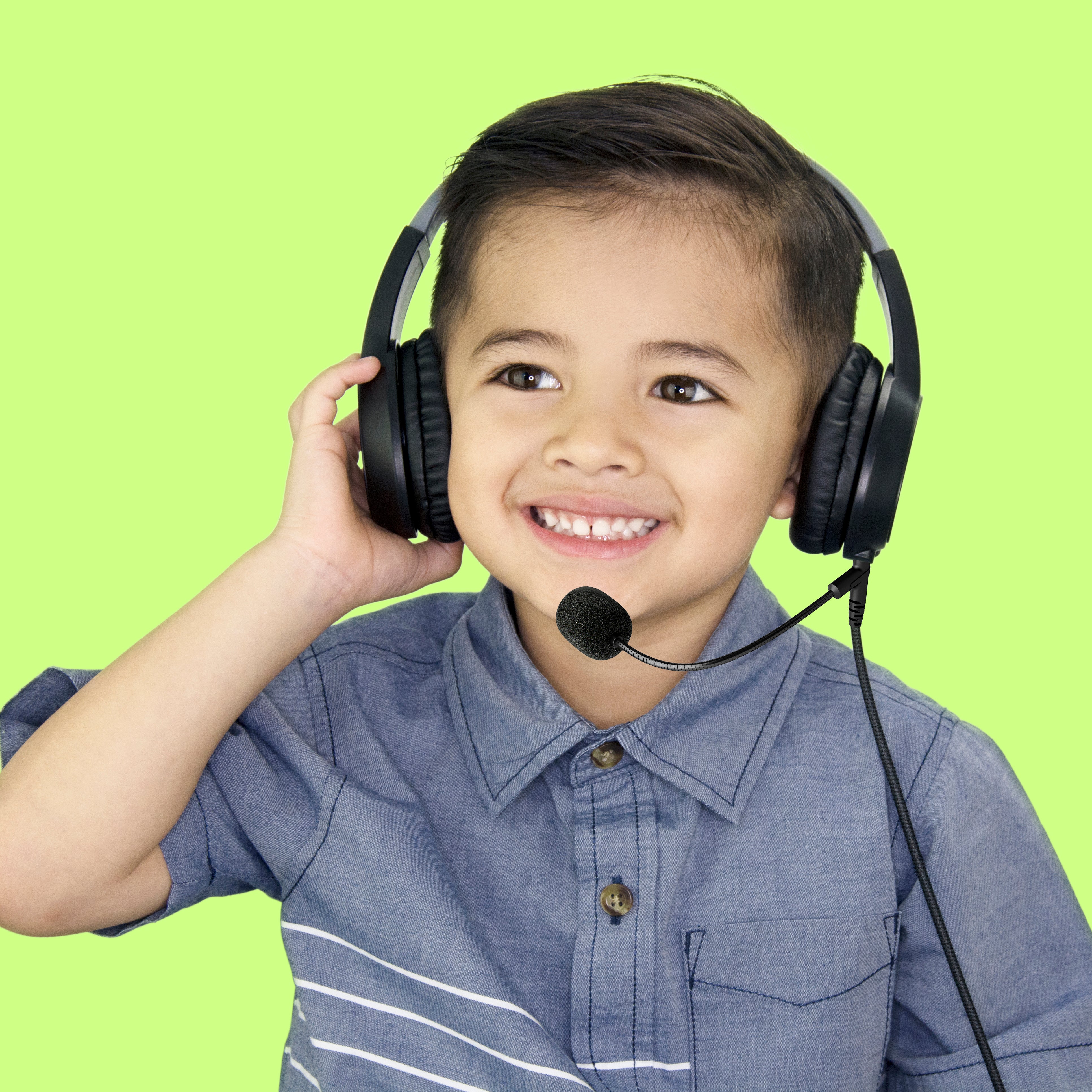 Young boy wearing a MEE audio KidJamz KJ35M with a microphone, smiling, and touching one earpiece, against a bright green background.