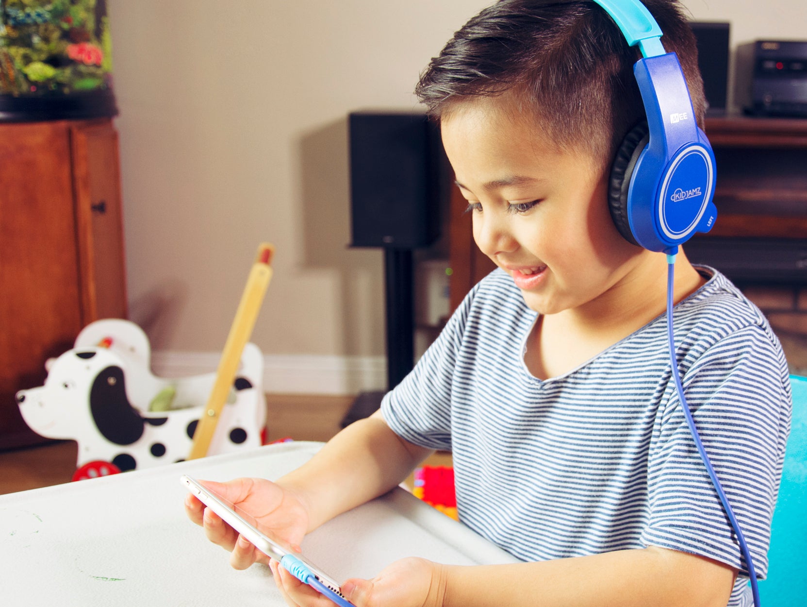 A smiling young boy wearing blue KJ35P KidJamz headphones, using a smartphone at a white table with a pencil and toy visible in the background.