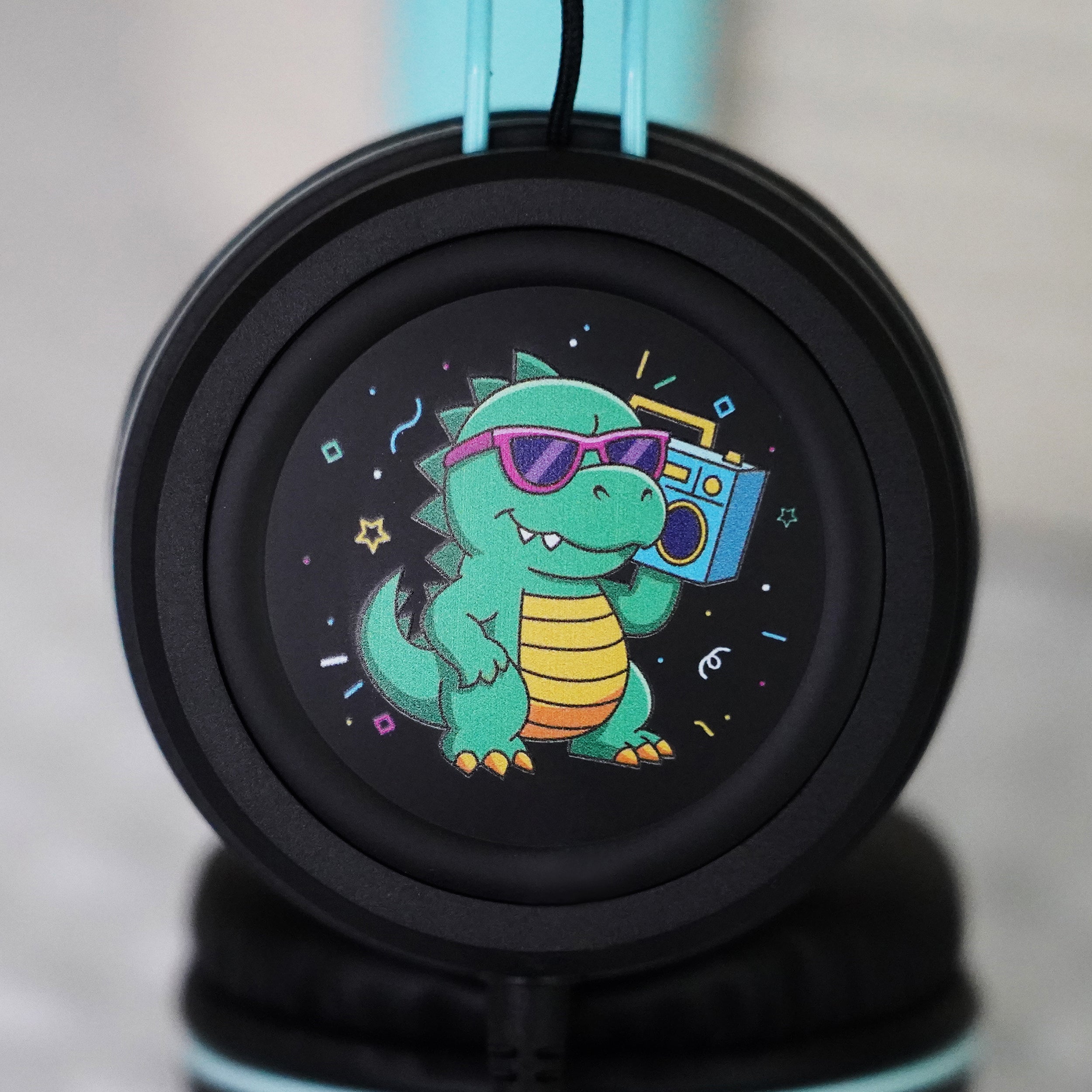 Black KidJamz KJ55AUX headphones faceplate with a cartoon Dinosaur design holding a speaker while in color on a light gray background