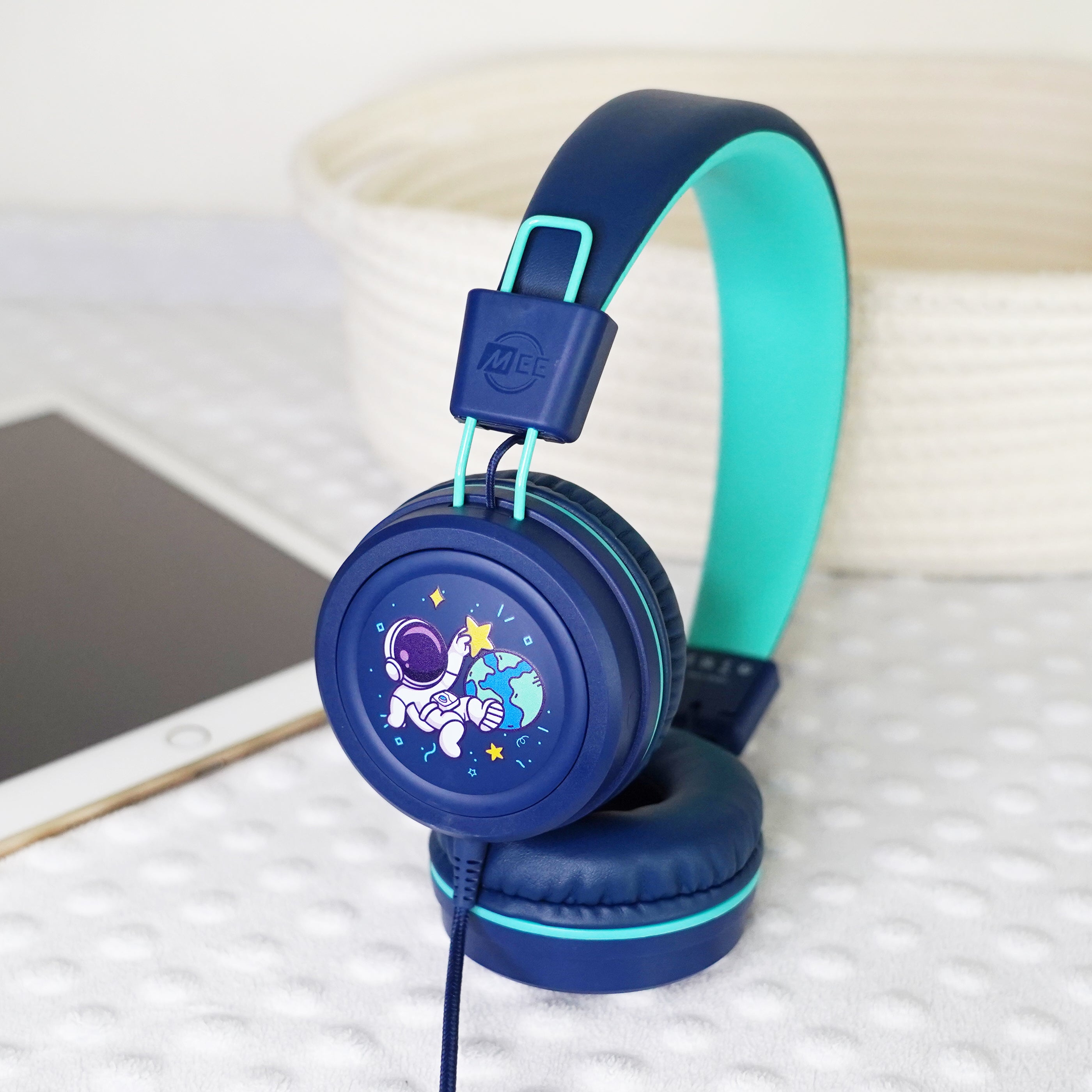 Blue KidJamz KJ55AUX headphones faceplate with a cartoon astronaut design holding a star while in outer space in color next to a tablet.