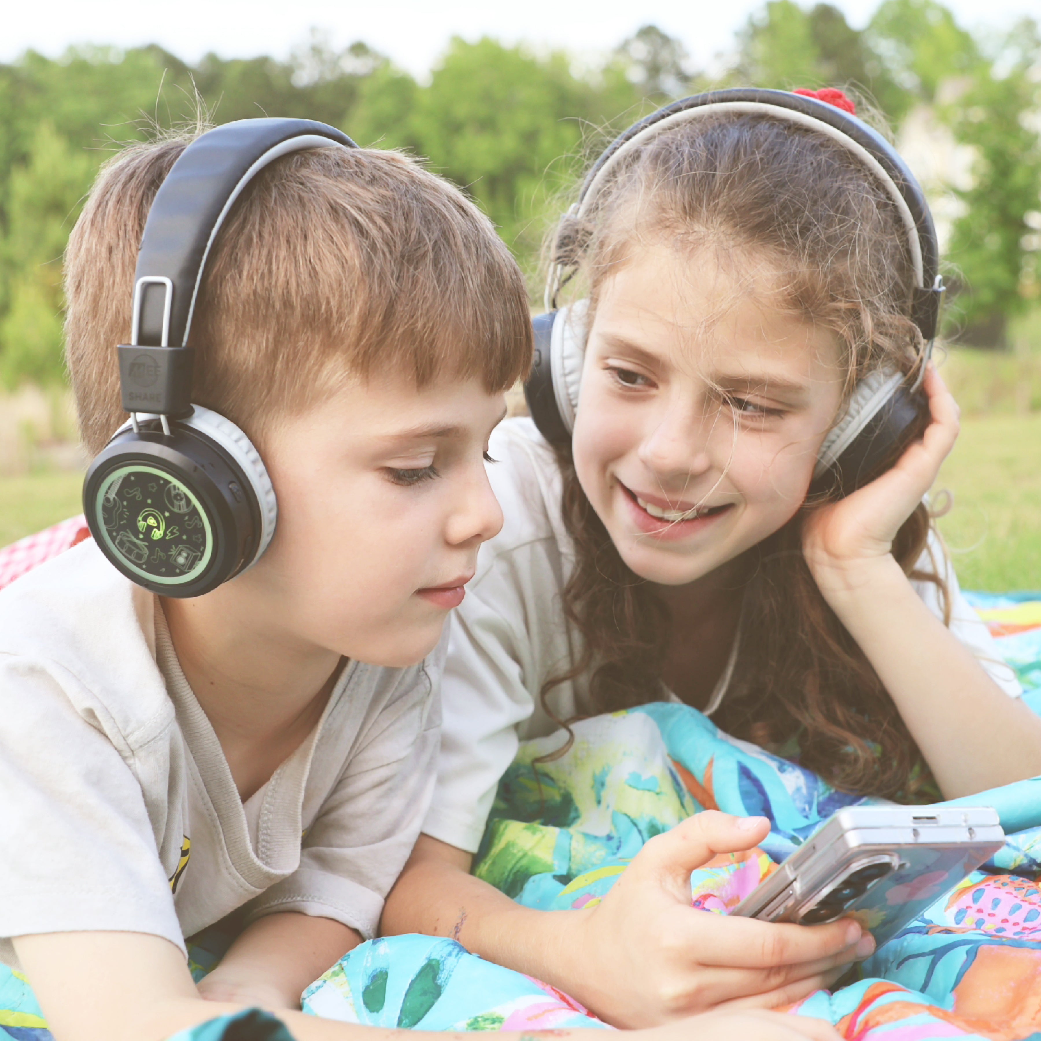 Two children wearing the MEE audio KidJamz Share Bluetooth Headphones for Kids with Wireless Audio Sharing in Sharing Mode that lets you easily link up to 1000 KidJamz Share together and stream audio from a single Bluetooth device - like a phone or tablet - to all of them at once, wirelessly!