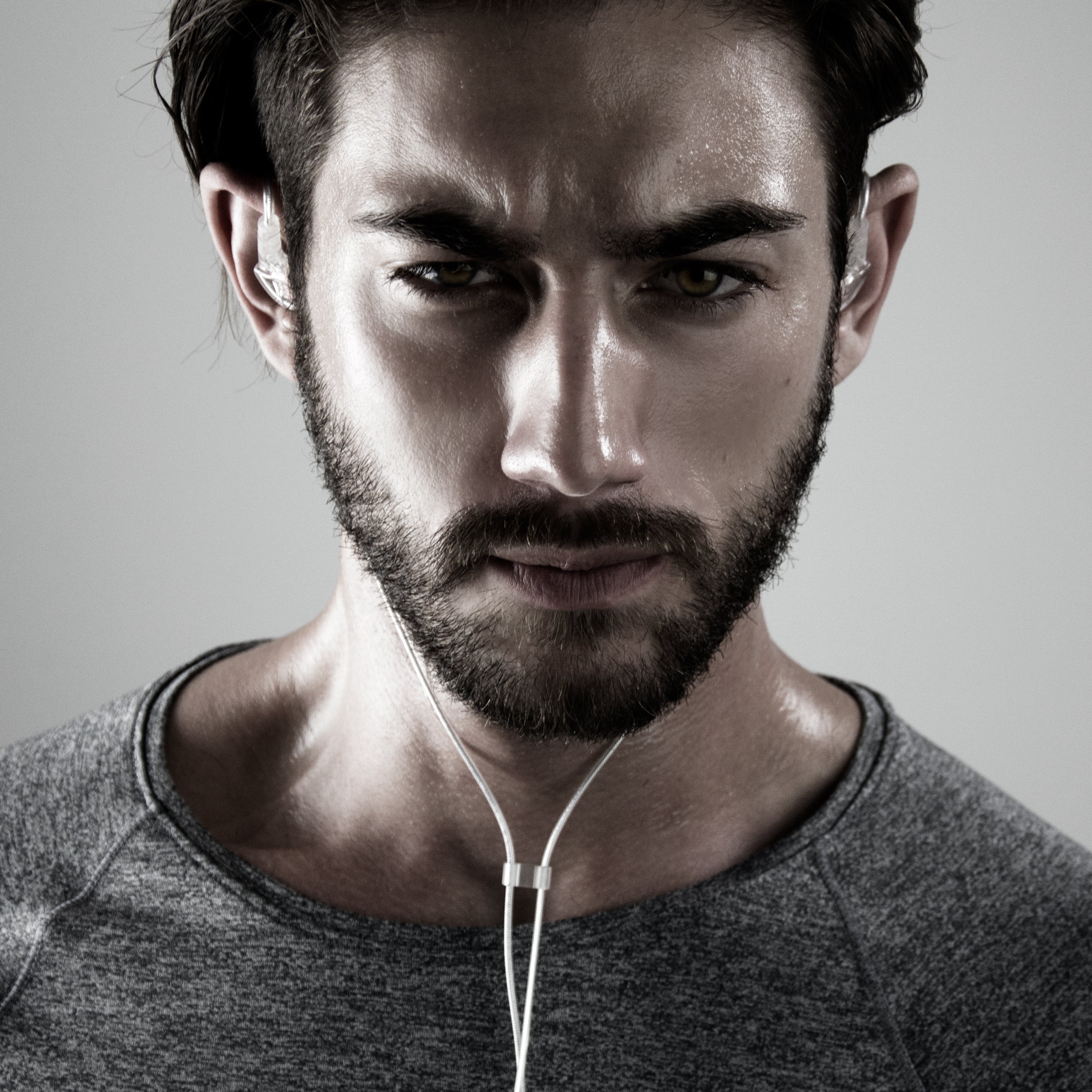 Close-up portrait of a man with a beard, intense gaze, and earphones, wearing a gray t-shirt on a neutral background.