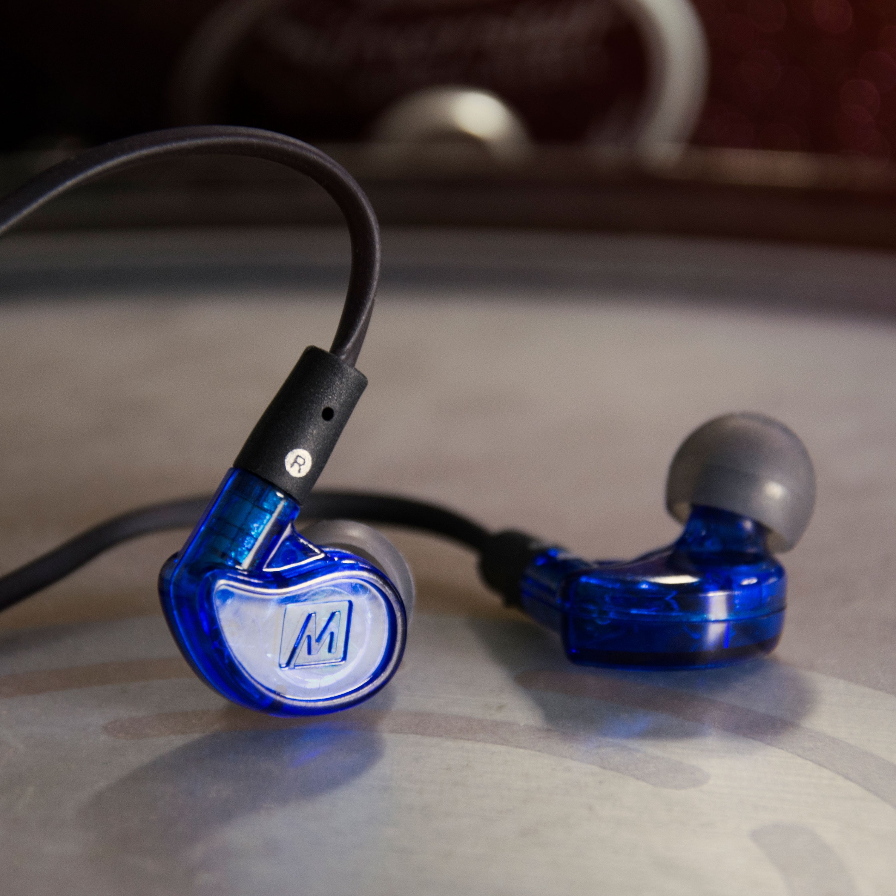 A pair of blue, transparent in-ear headphones with a visible logo on the sides, resting on a gray surface against a softly blurred background.