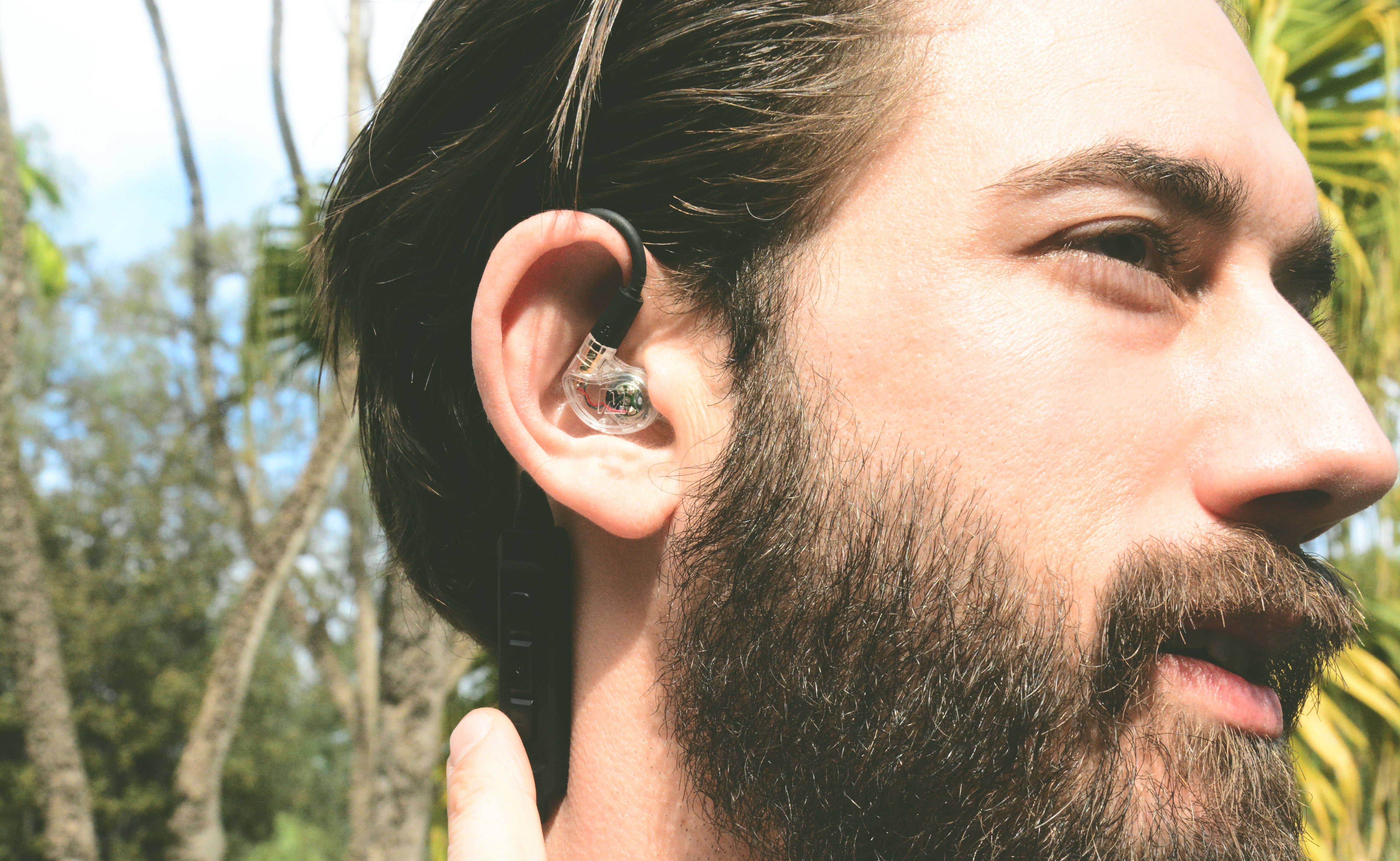 Man with a beard wearing MEE audio M6 PRO Musicians' In-Ear Monitors Wired + Wireless Combo Pack: Includes IEMs, 2 Audio Cables and BT3 Hi-Res Bluetooth Audio Adapter w/aptX, LDAC, Auracast Support, & Built-In Headset (Clear) outdoors