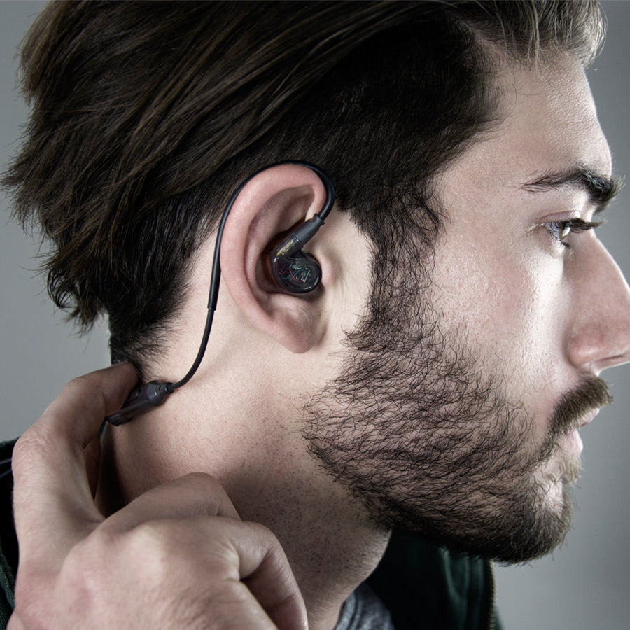 A close-up photo of a young man from the side, putting a black wireless in-ear monitor into his right ear. he has a beard and his focus seems directed towards fitting the earphone comfortably.