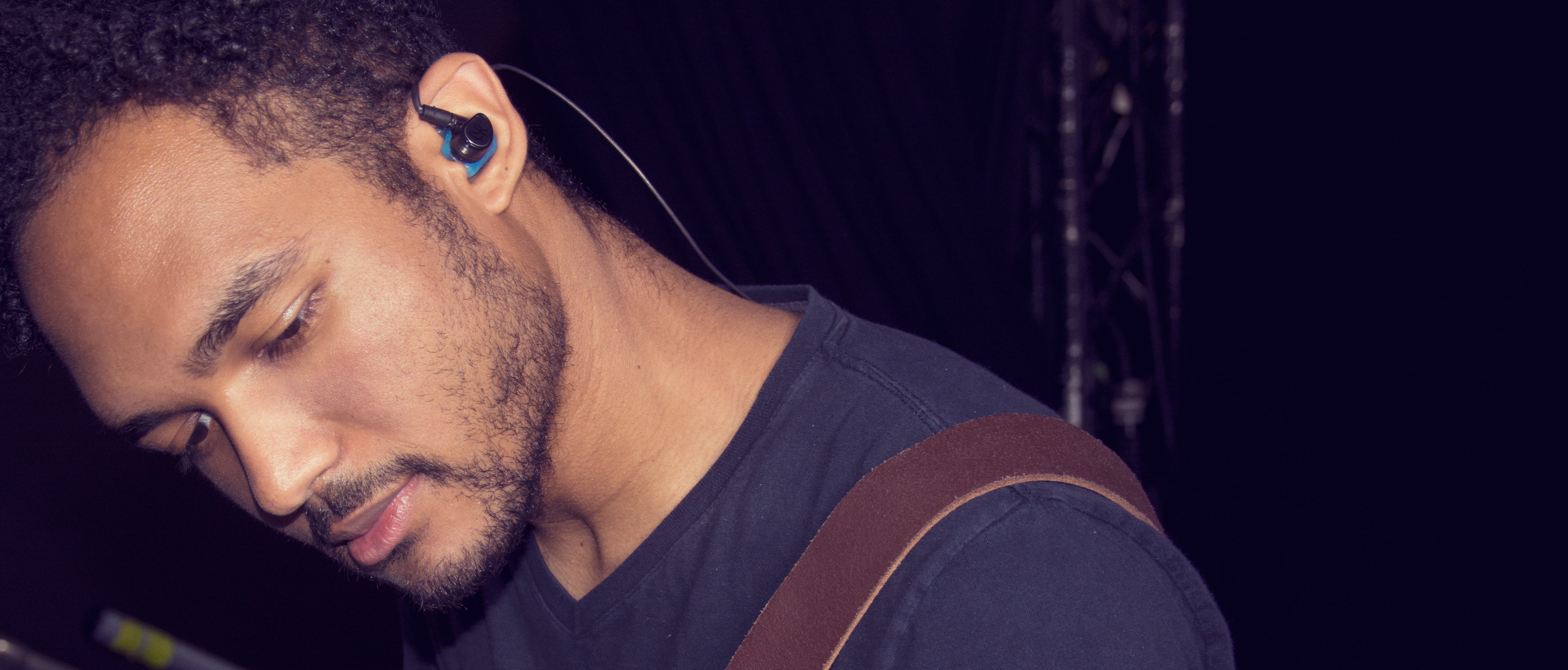 A close-up side view of a young man wearing a wireless earphone and carrying a guitar strap, focused on his work in a dimly lit setting.