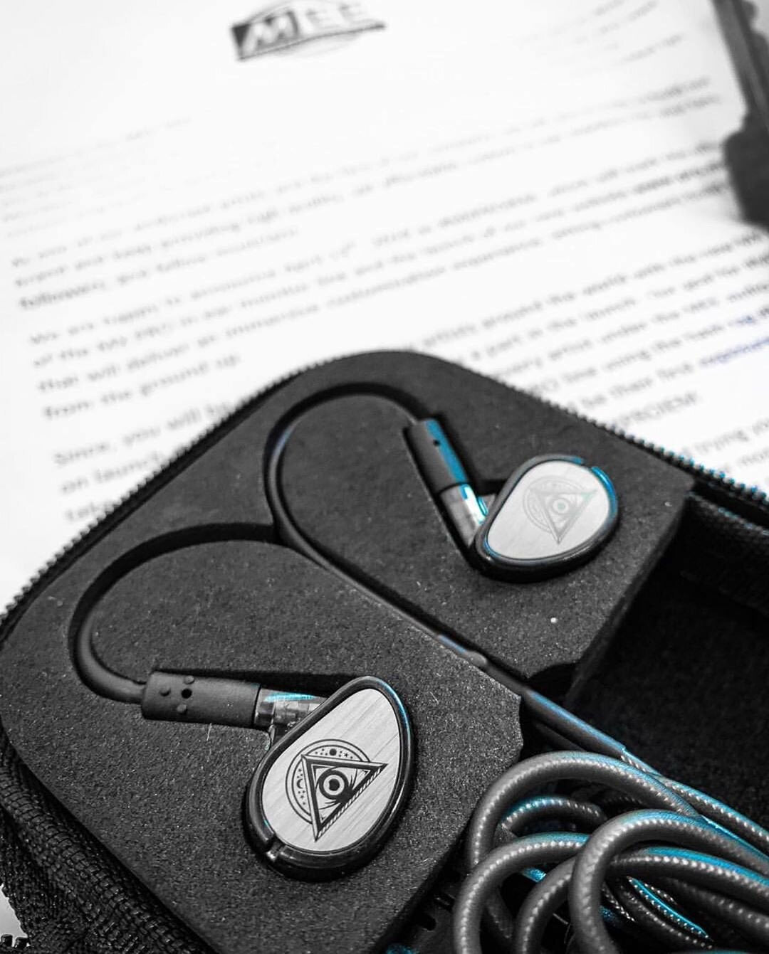 A pair of high-end in-ear headphones with metallic design and balanced armature drivers, sitting in a foam insert of its storage box, contrasted against a blurred document in the background. color highlights in blue and tones in grayscale.