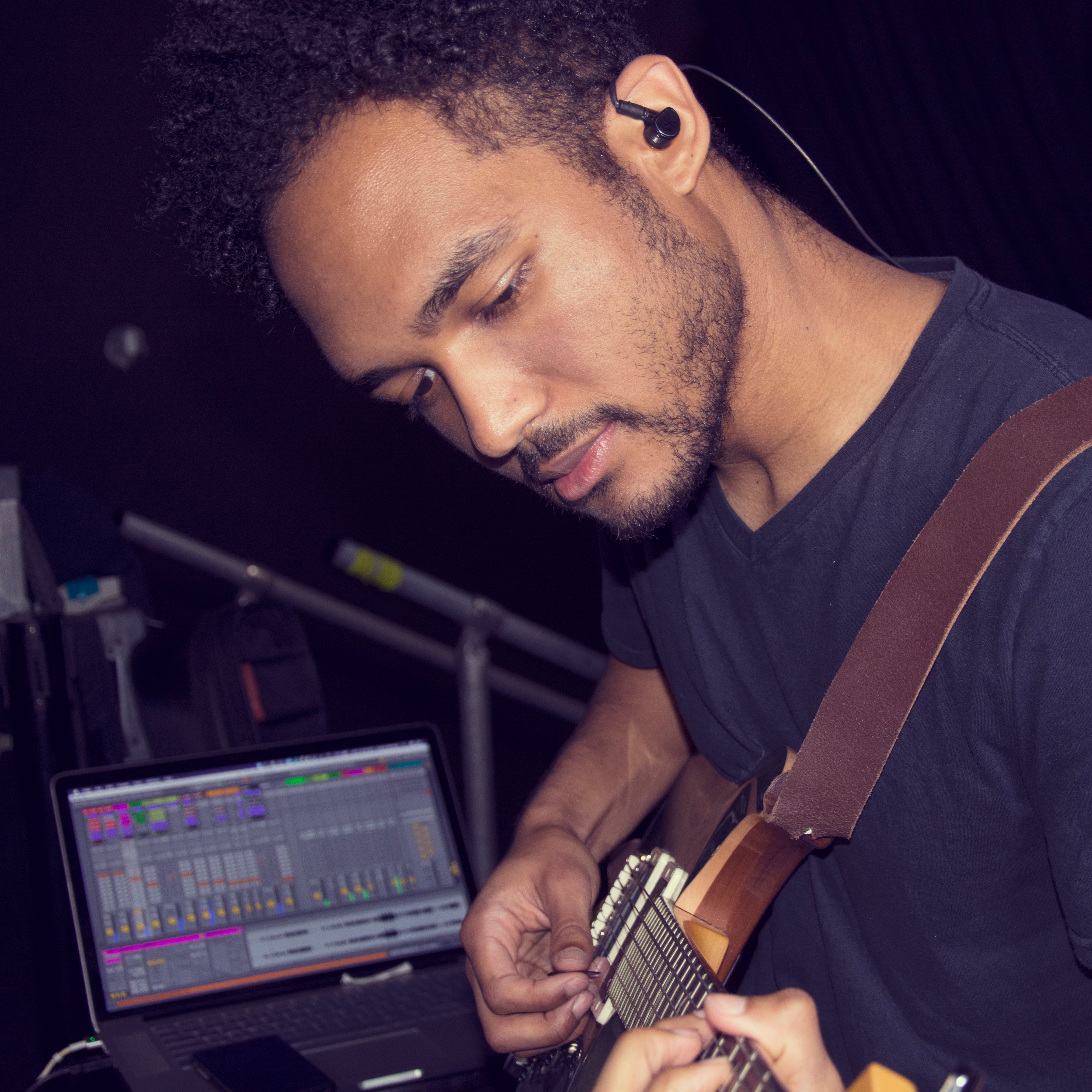 A young man tuning a guitar while looking at a computer screen with music production software open, in a dimly lit room.