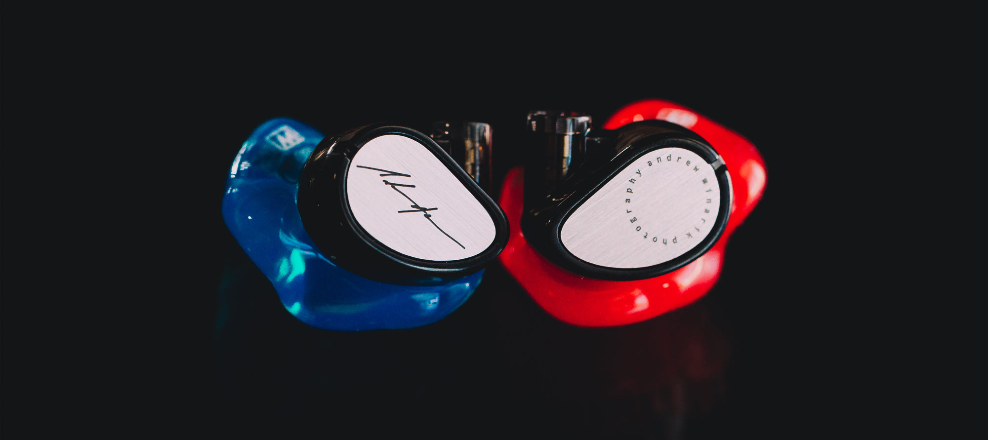 Two high-end in-ear monitors with custom ear molds, one blue and the other red, featuring metallic faceplates with stylish logos, displayed against a dark reflective background.