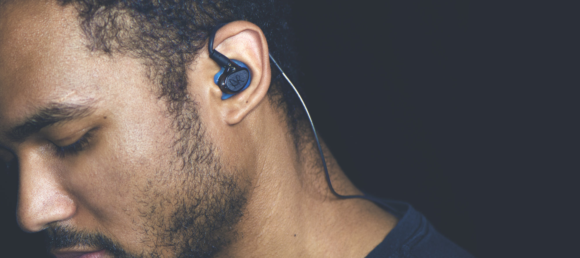 A close-up of a man's side profile showing a wireless earbud in his ear, with his face partially lit against a dark background.