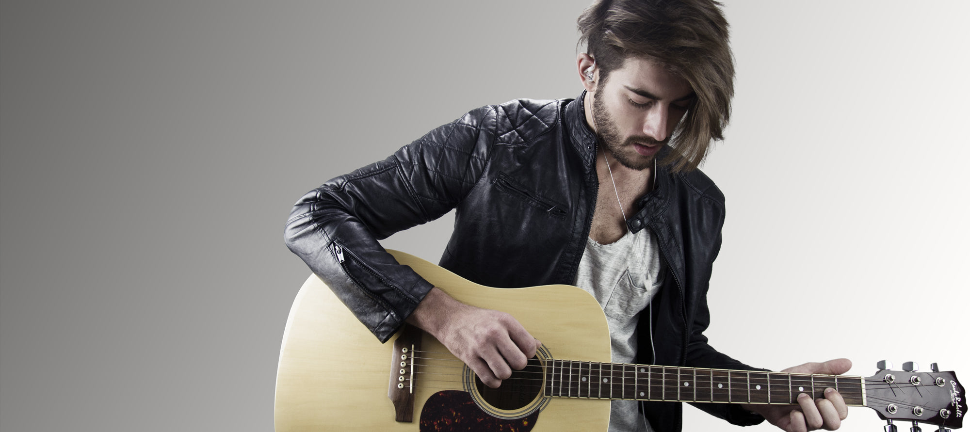 A stylish young man with a beard, wearing a leather jacket, plays an acoustic guitar against a light gray background.