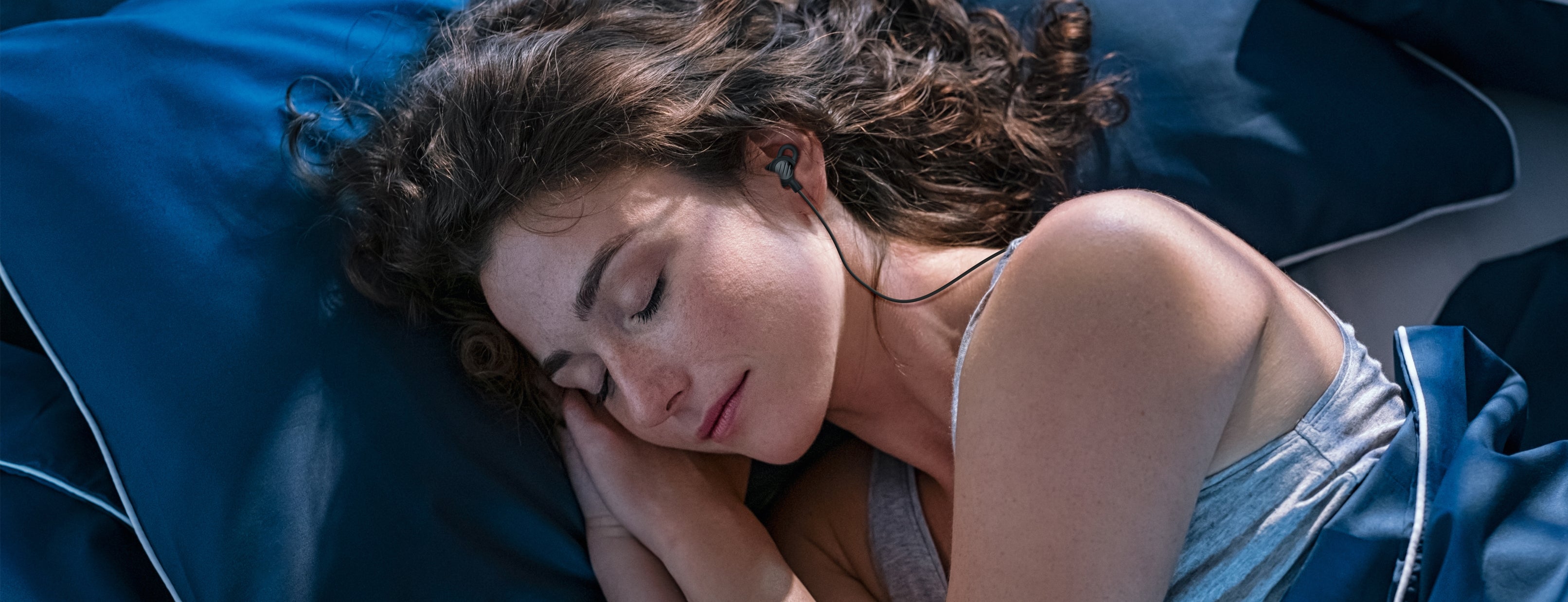 Woman sleeping peacefully on a blue pillow with the MEE audio SleepTones earbuds.