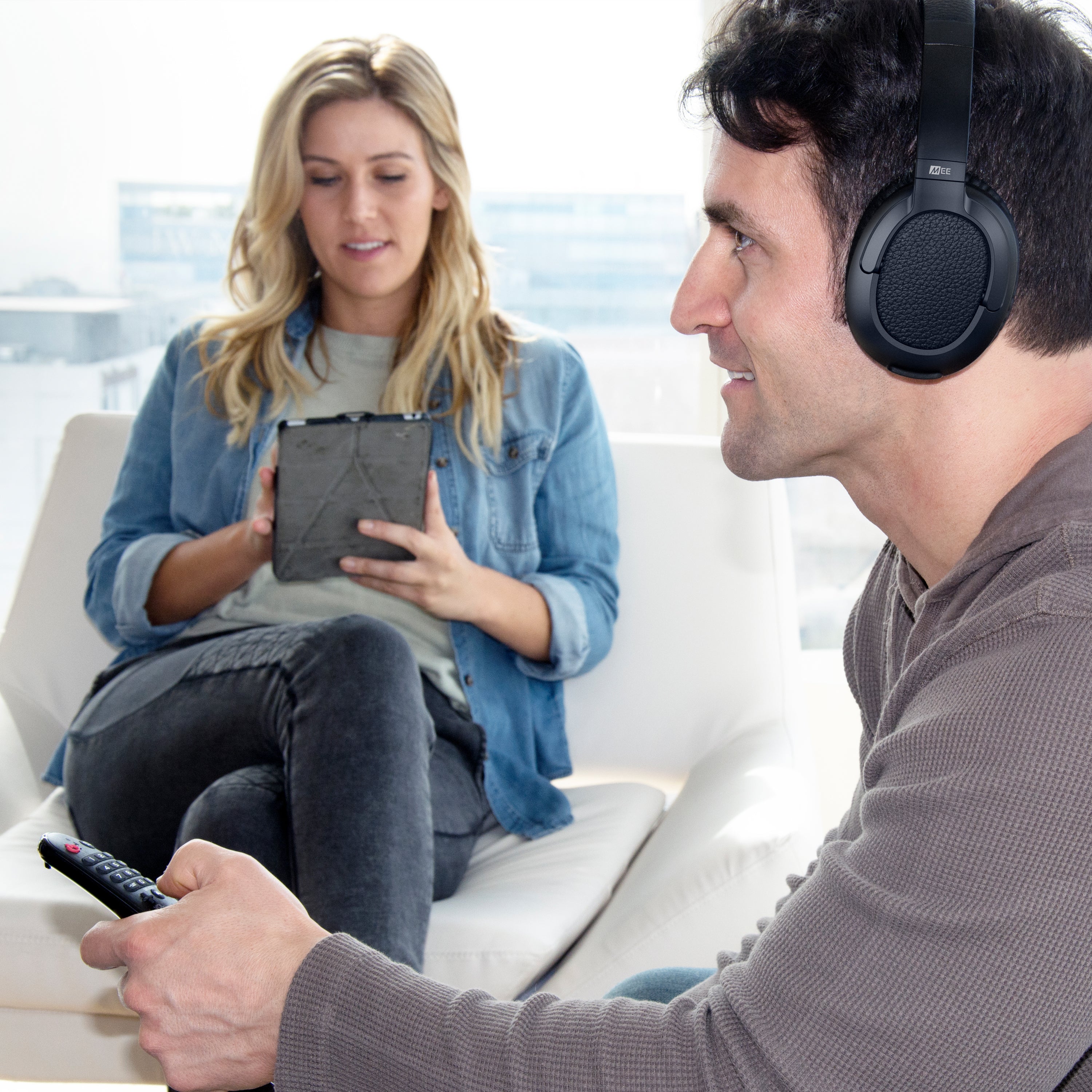 A man wearing headphones holds a remote, looking at a woman seated nearby who is focused on a tablet, both sitting on a white couch in a brightly lit room.