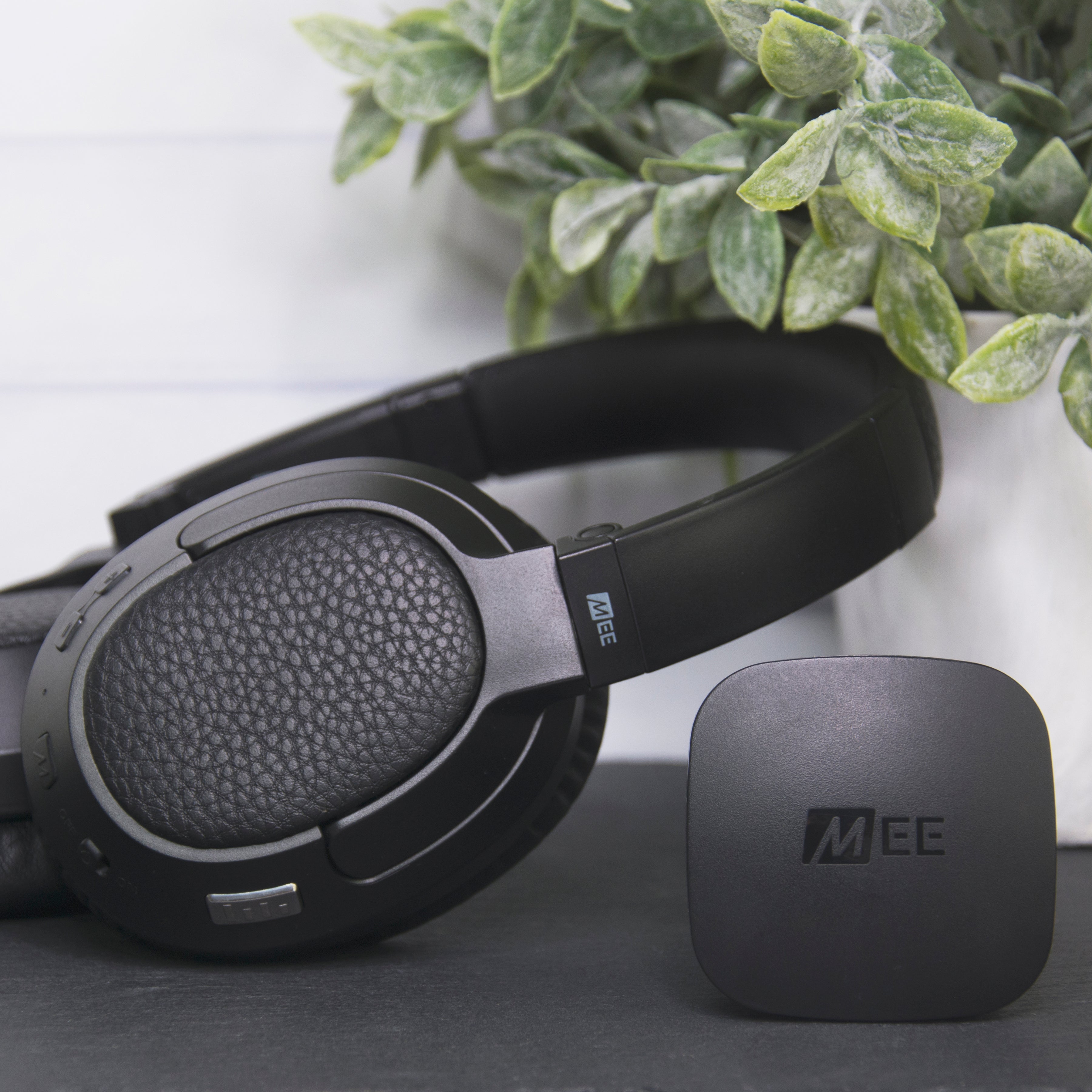 A close-up of a pair of over-ear headphones with textured ear cups, next to a small black electronic device, against a backdrop of a green plant and white wooden slats.