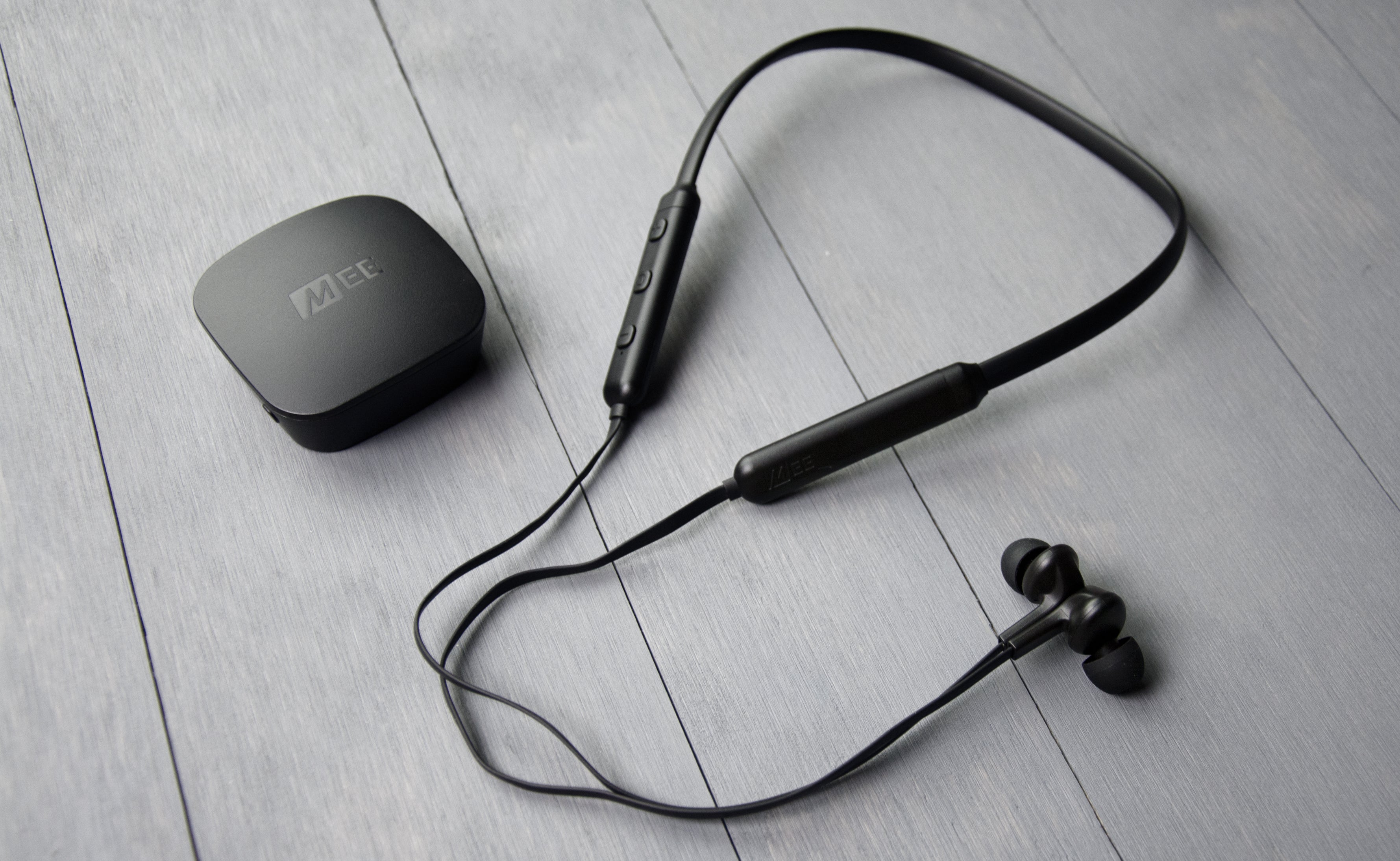 Wireless neckband earphones with inline controls and magnetic earbuds next to their charging case, resting on a gray wooden surface.