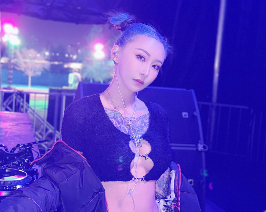 A woman with blue-tinted hair styled in buns, wearing a black crop top and open jacket, stands in front of a dj setup with colorful stage lights in the background.