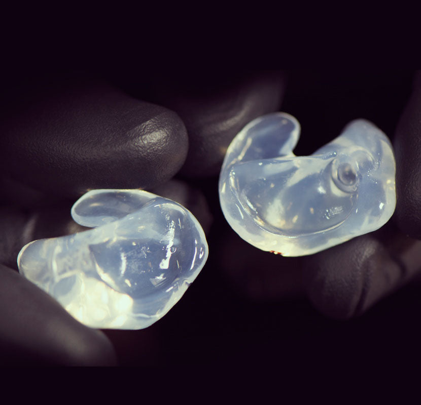 Two hands holding a pair of translucent, custom-molded earpieces against a dark background. the earpieces are illuminated, emphasizing their detailed shapes.