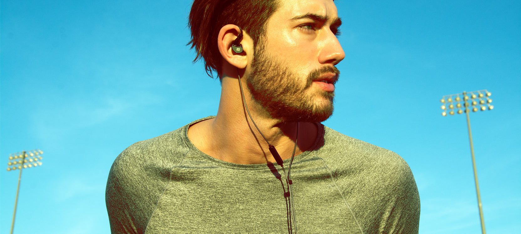 Young man with a beard, wearing earphones and a grey t-shirt, looks to the side under a bright blue sky with stadium lights in the background.