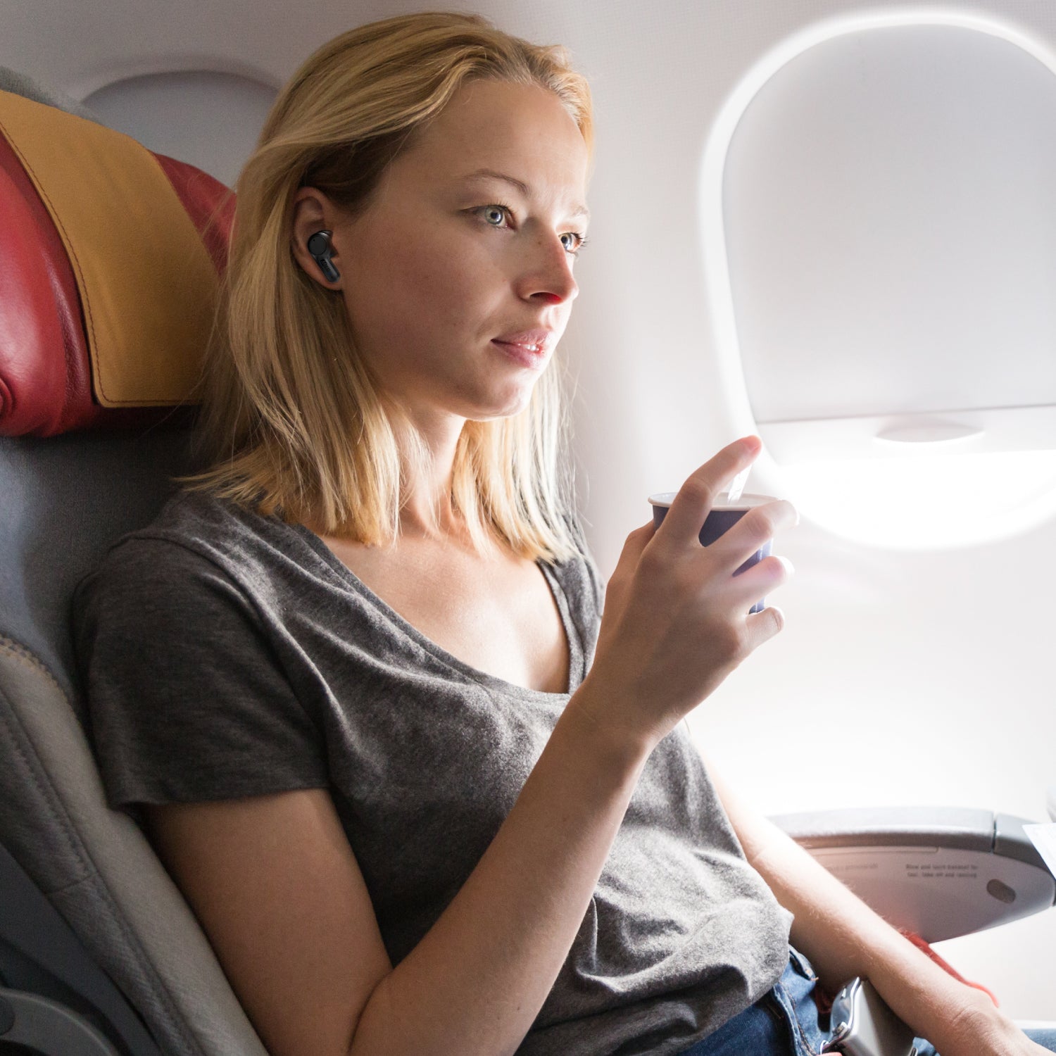 A woman with blonde hair sitting by an airplane window, wearing a gray t-shirt and using her smartphone, lit by natural light from outside.