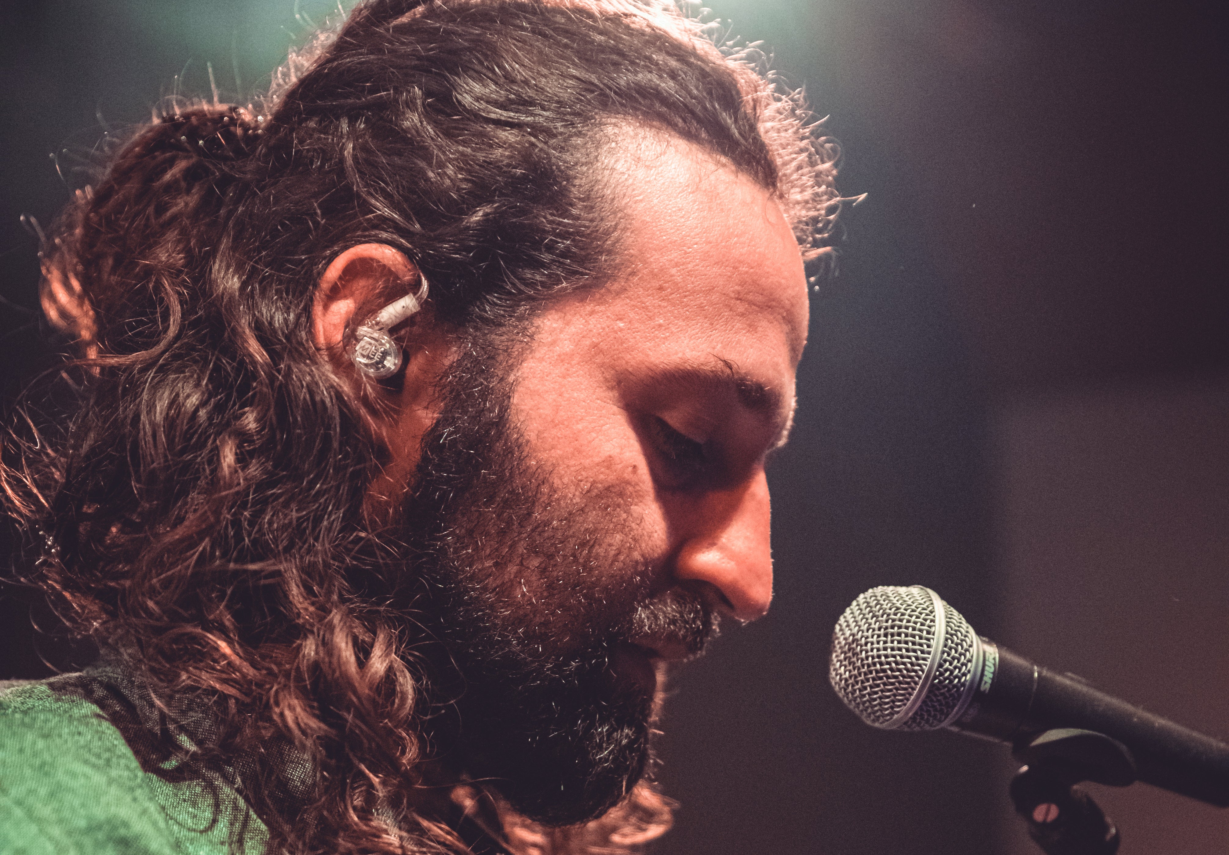 A close-up side view of a male singer with long curly hair singing into a microphone on stage, showcasing a focused expression under warm stage lighting.