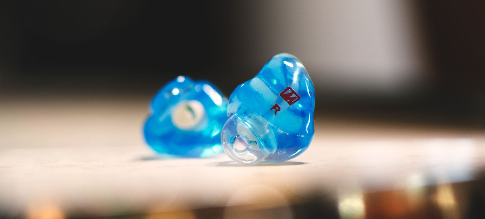 Two custom-molded, blue in-ear monitors with a "r" marking on one, focused closely on a blurry wooden surface.