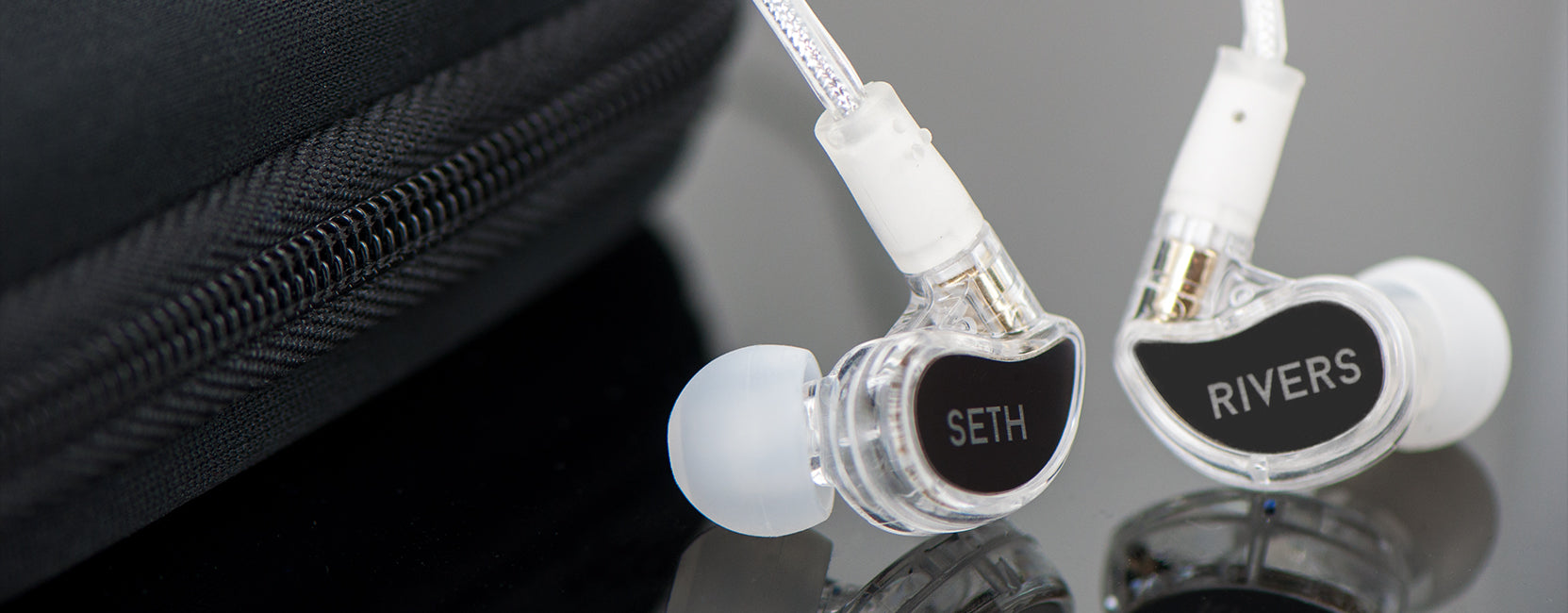 Two custom in-ear monitors with clear casings labeled "seth" and "rivers" placed in front of a black case on a reflective surface.