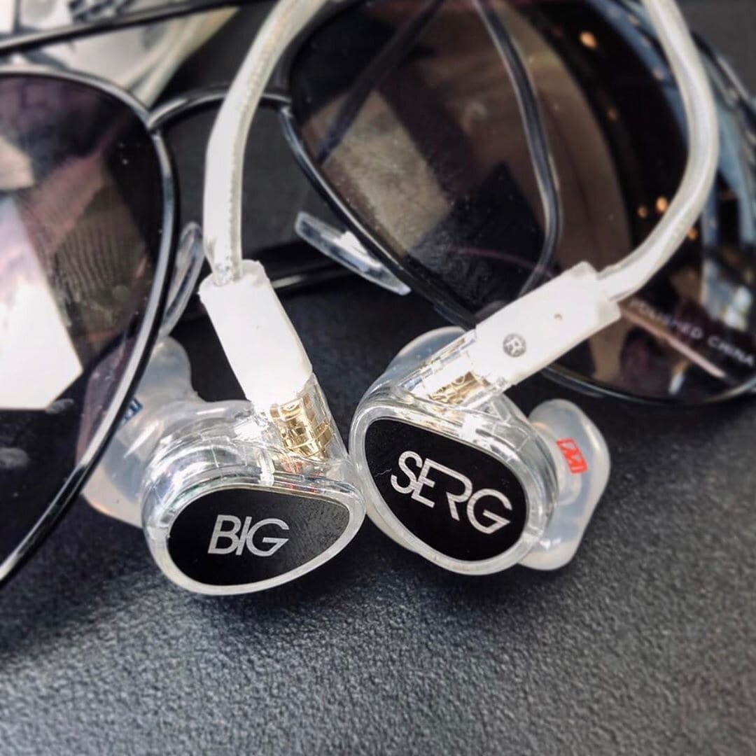 A close-up of two custom in-ear monitors with transparent casings and engraved logos, "big" and "srg," placed next to a pair of overlapping sunglasses on a dark surface.