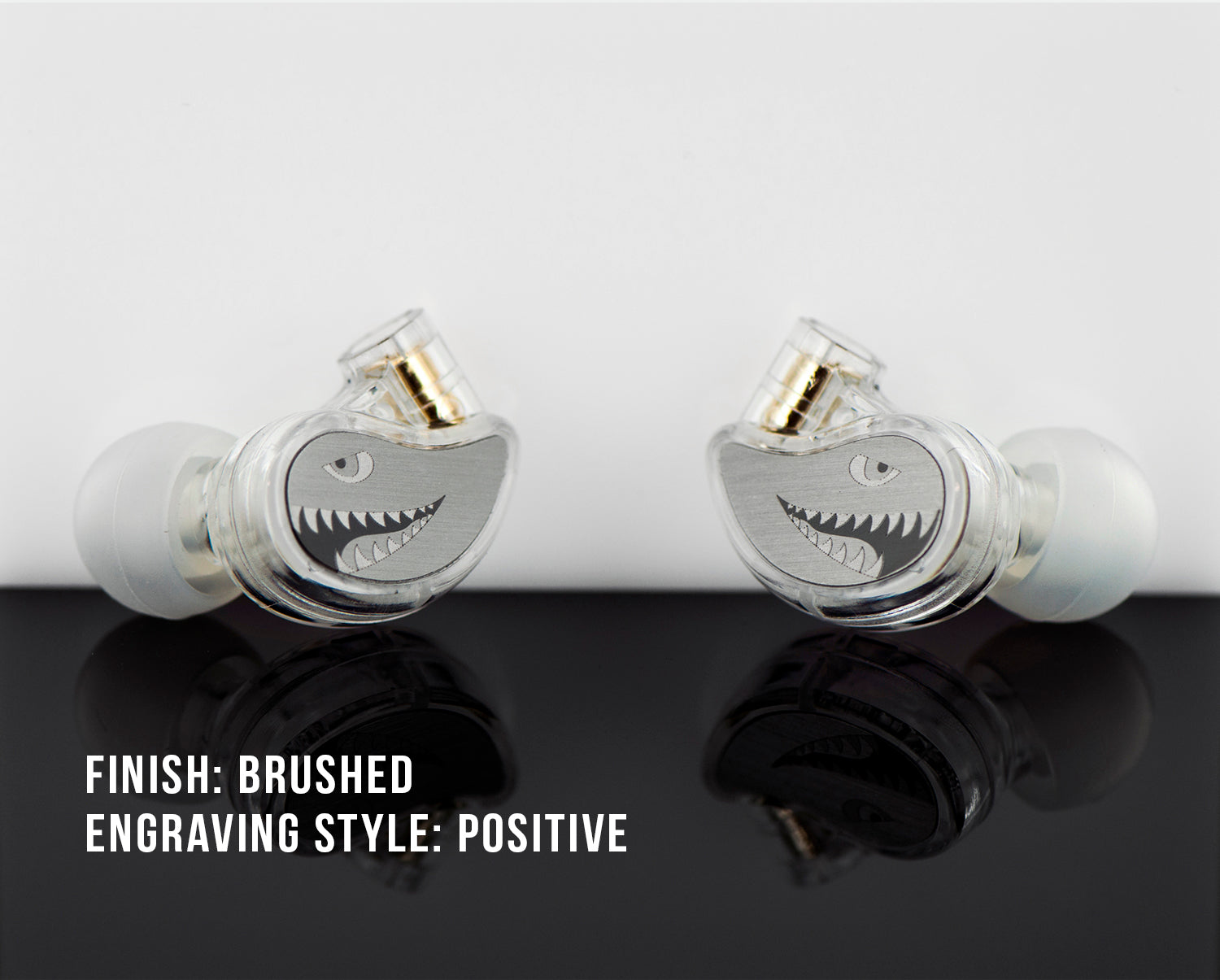 Two in-ear monitors with a brushed finish and shark face engravings, displayed on a reflective surface with text labels "finish: brushed" and "engraving style: positive".