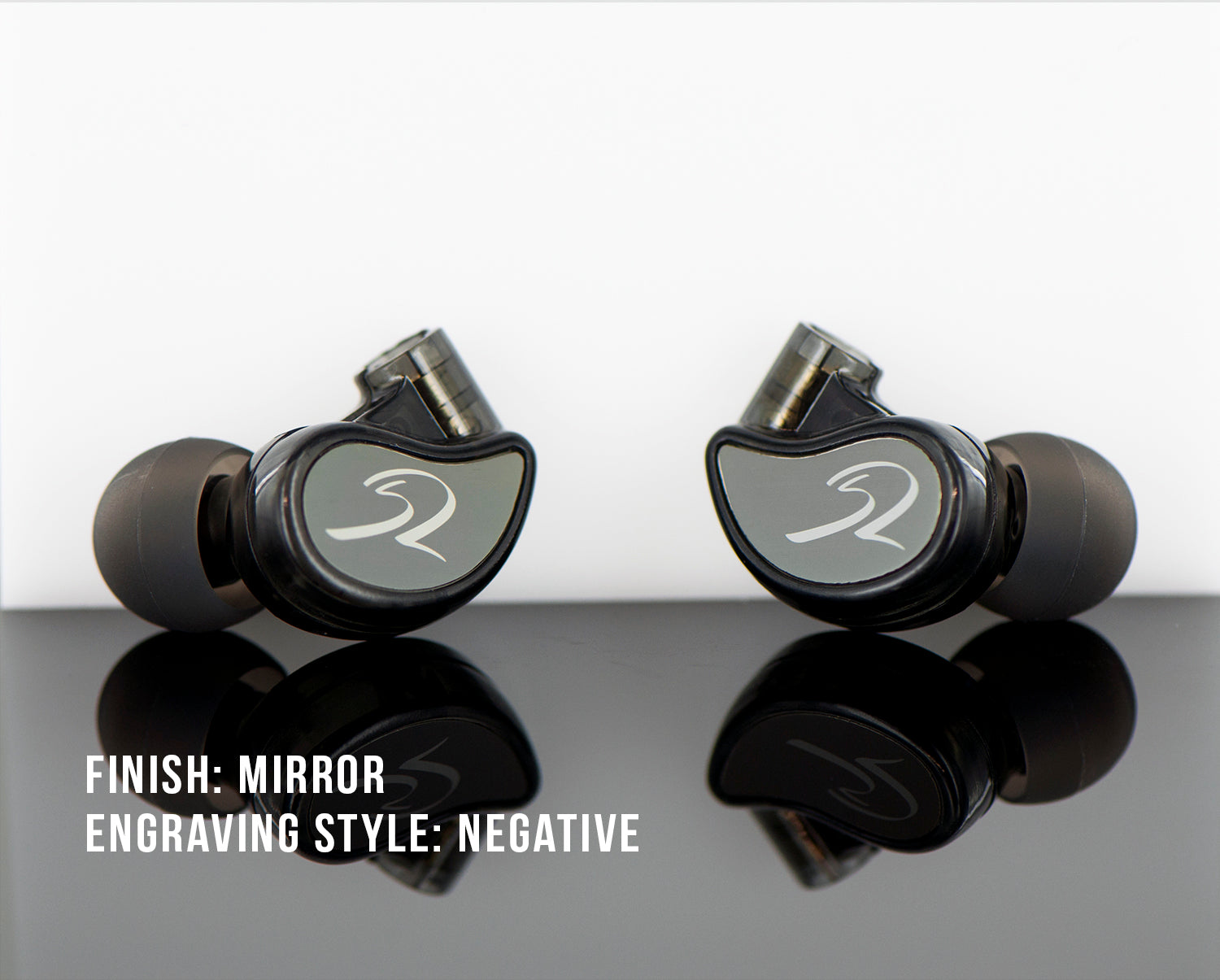 Two black in-ear headphones with a mirrored finish and a white logo, displayed on a reflective surface, with the text "finish: mirror engraving style: negative" below.