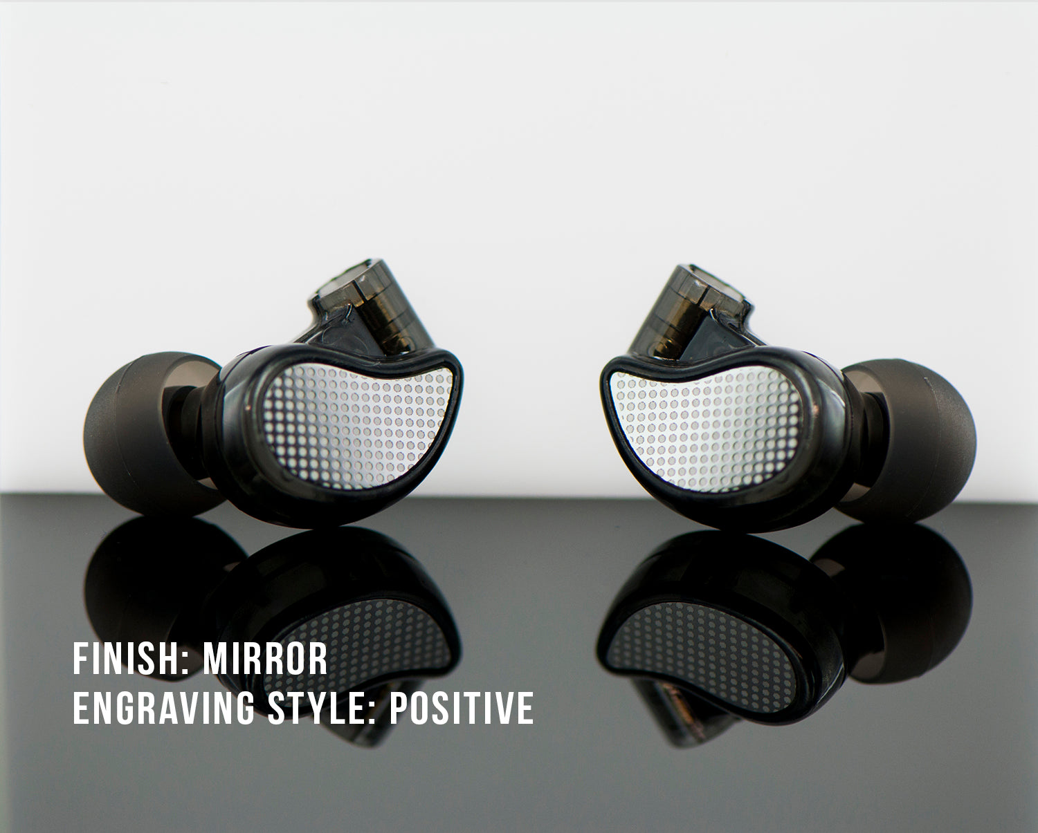 Two high-end in-ear headphones with a mirror finish and positive engraving style on their outer shells, reflected on a glossy surface, against a light grey background.